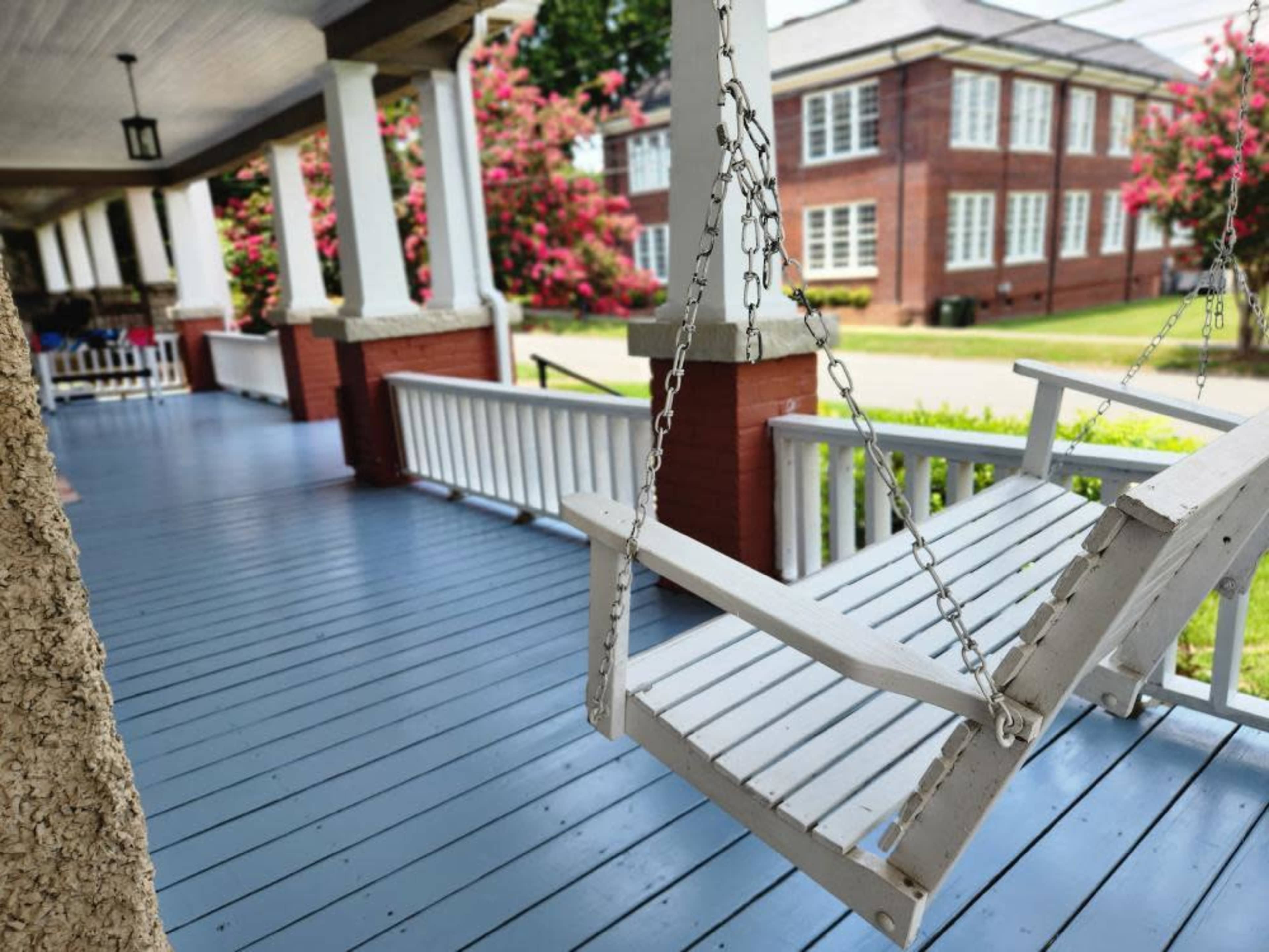 A white swing hangs from a porch overlooking a grassy area and a brick building in the background.