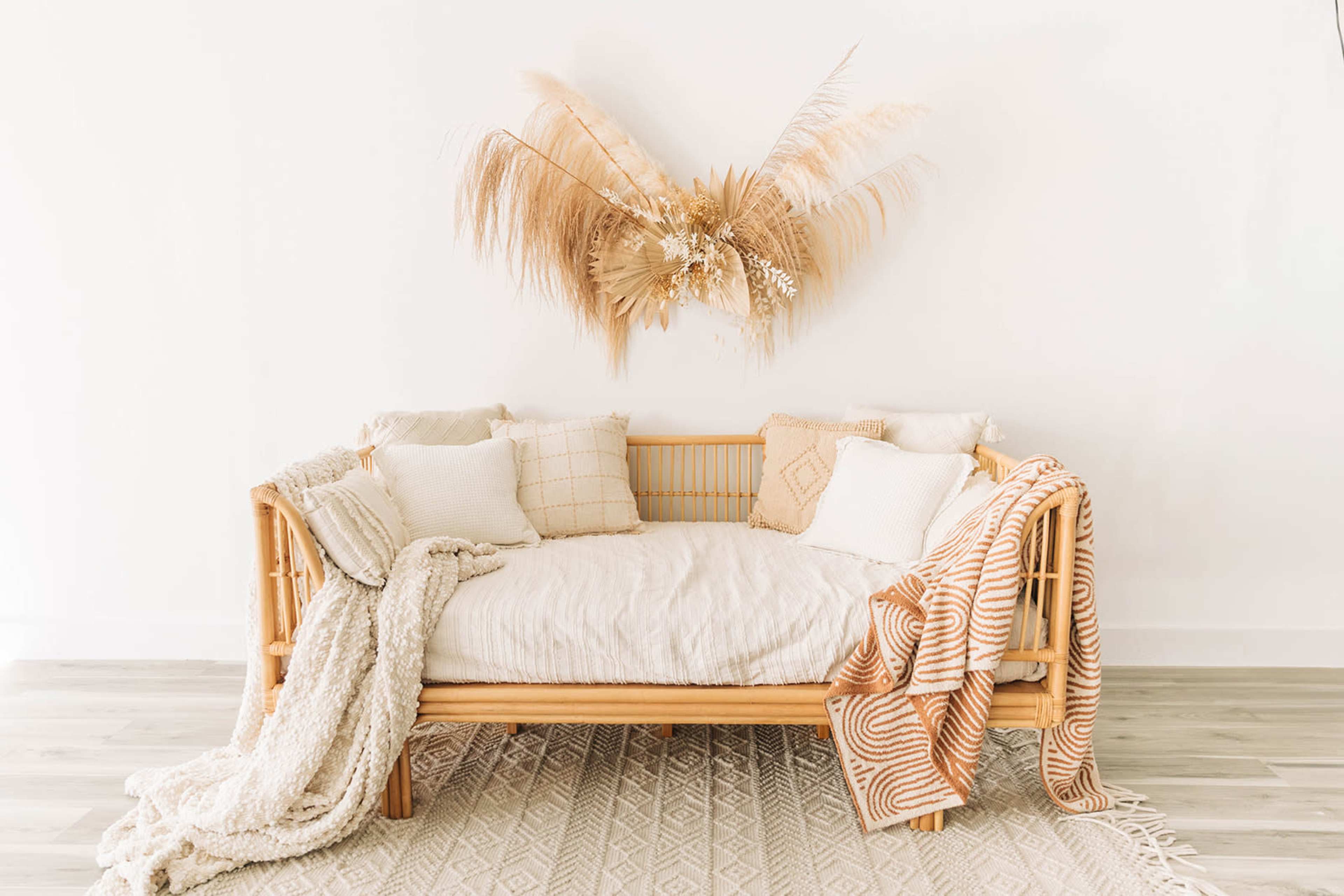 A rattan daybed adorned with various textured pillows and blankets is positioned against a white wall, featuring dried floral decor above it.