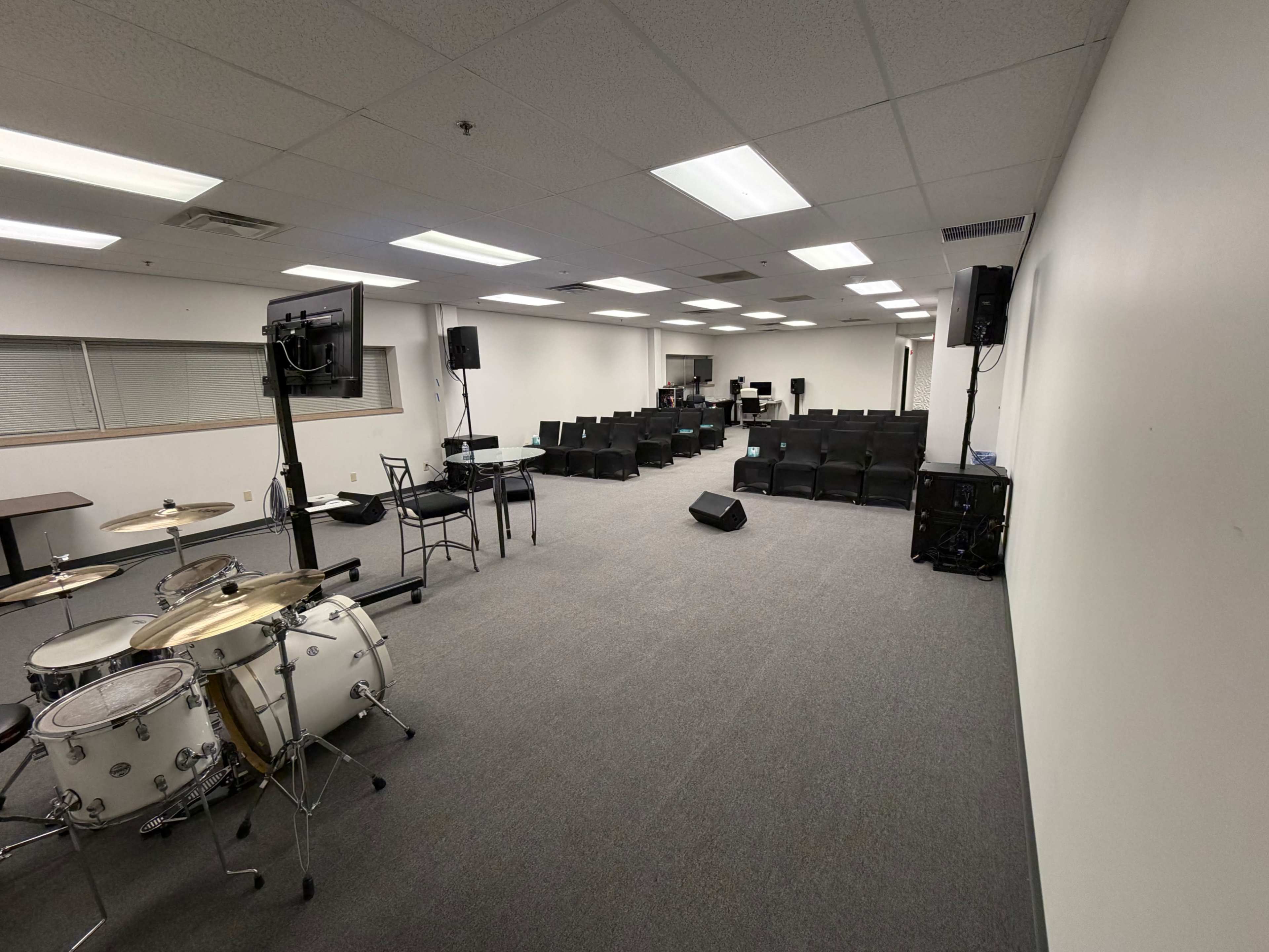 The image shows a large room set up for an event, featuring a drum set on one side and rows of black chairs arranged for an audience.