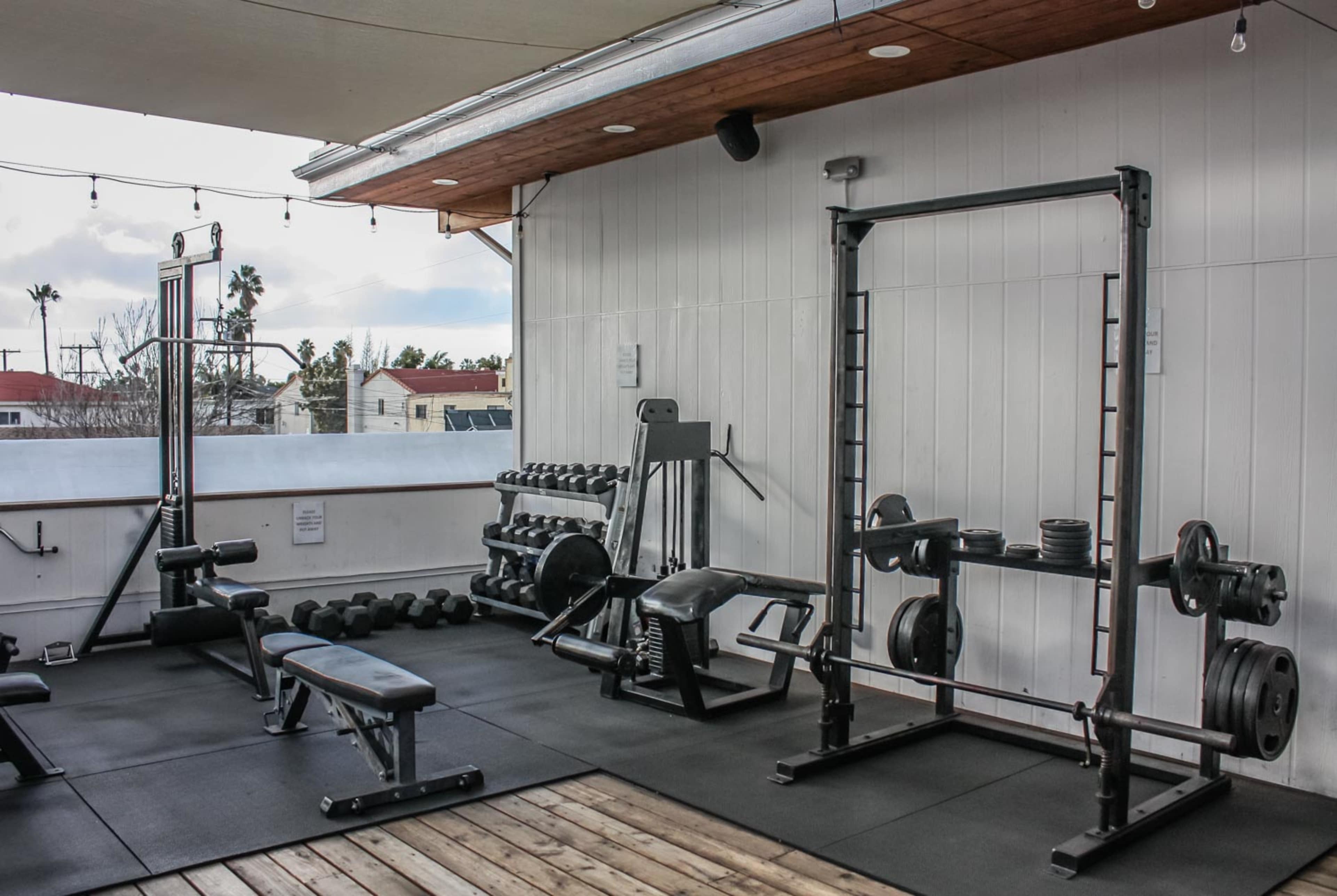 An outdoor gym area featuring various weightlifting equipment, including a bench press, a squat rack, and free weights on a balcony.