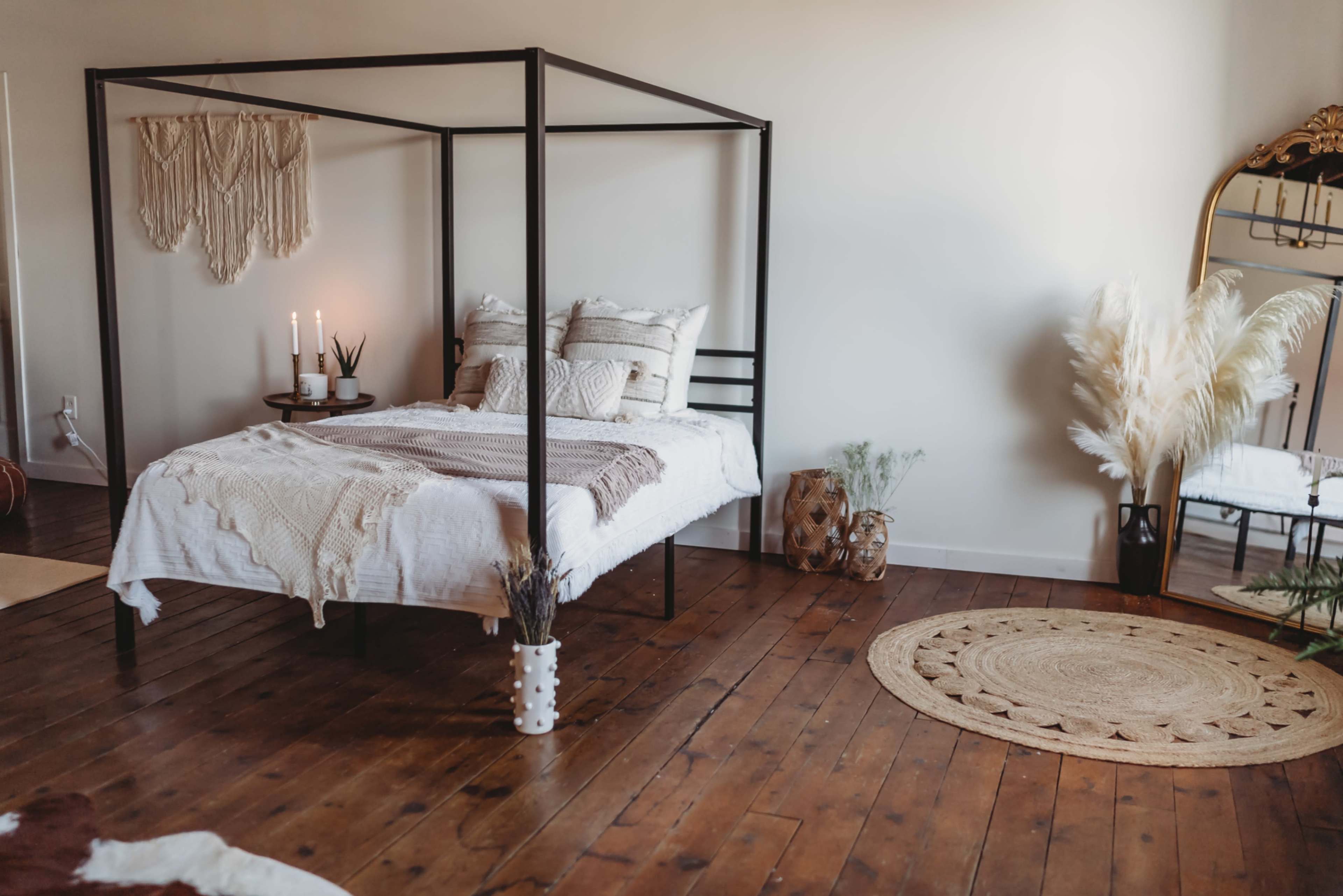 A four-poster bed with a white comforter is centered in a cozy room featuring wooden floors and a large mirror.