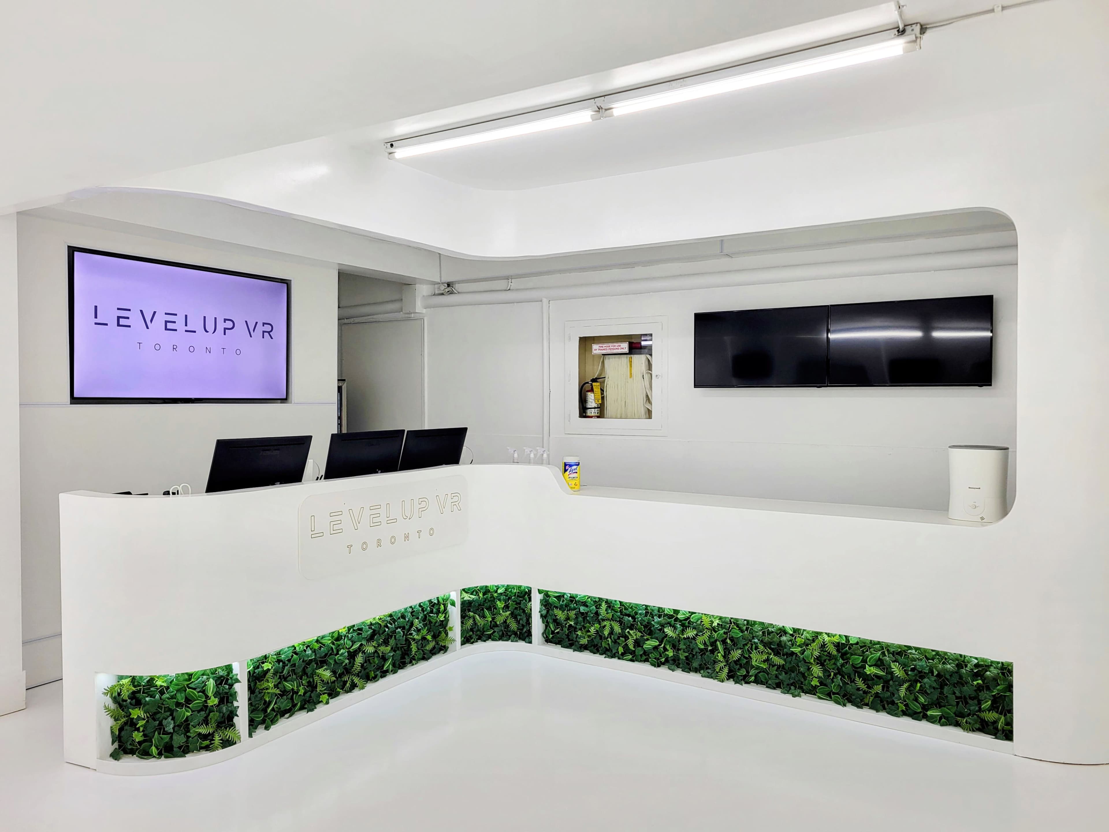 The image shows a modern reception area for Level Up VR in Toronto, featuring a sleek white counter with greenery at the base and a large screen displaying the company name.