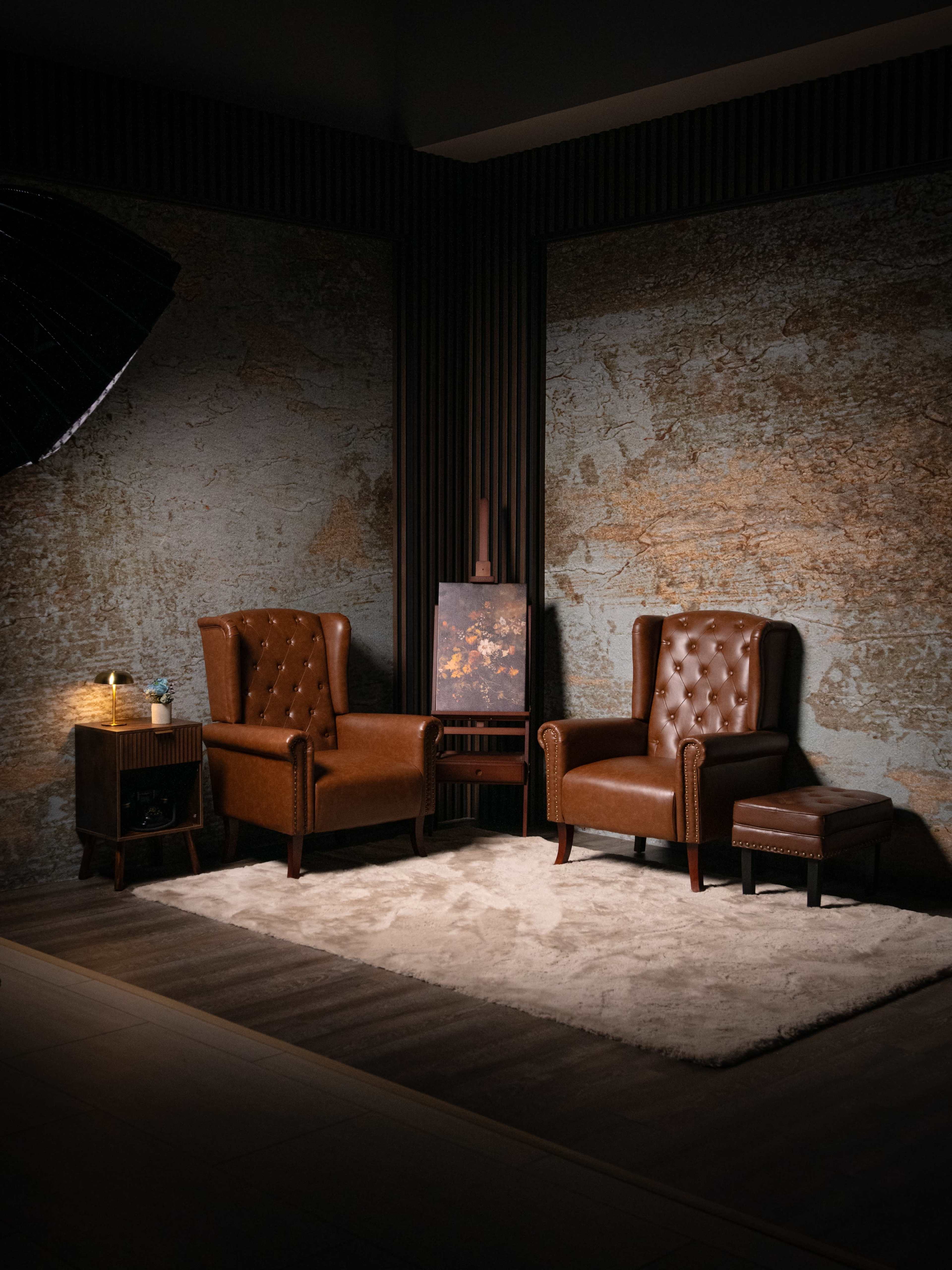 Two brown leather armchairs are positioned on a rug under soft lighting in a corner with textured walls.