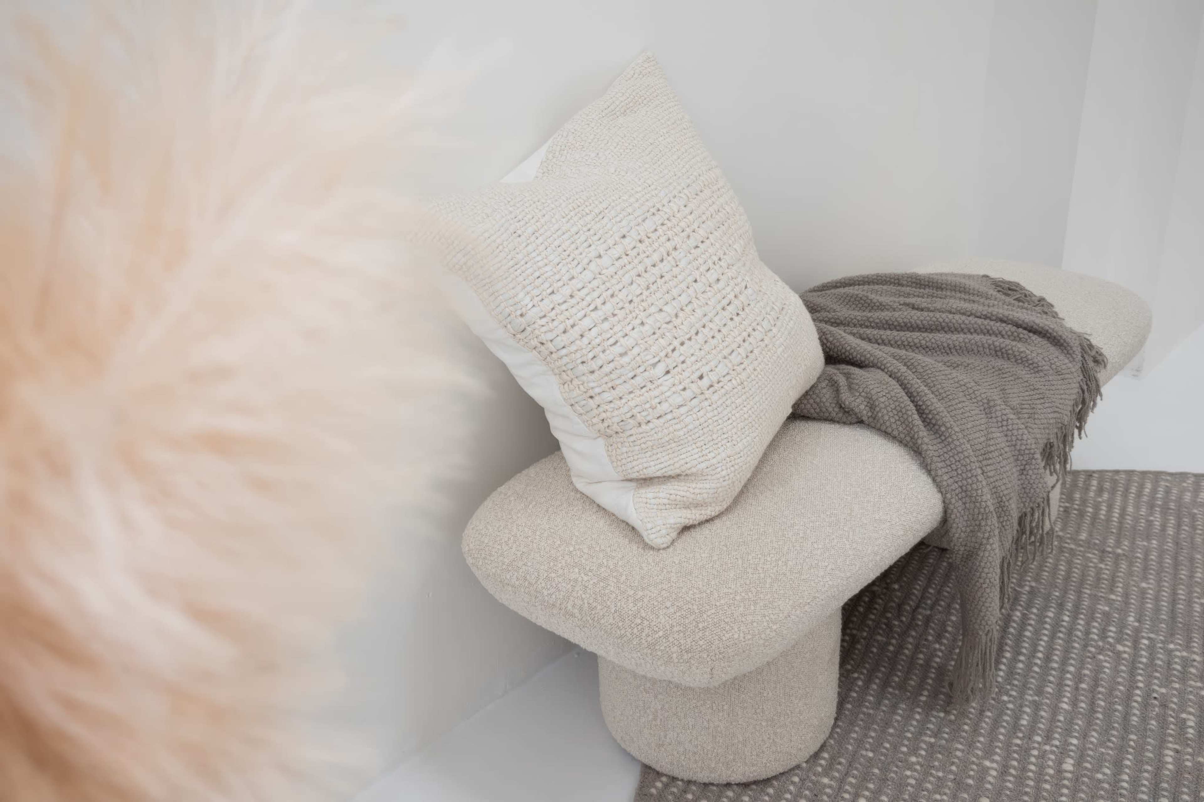 A textured pillow and a gray blanket are draped over a beige, curved bench against a light-colored wall.