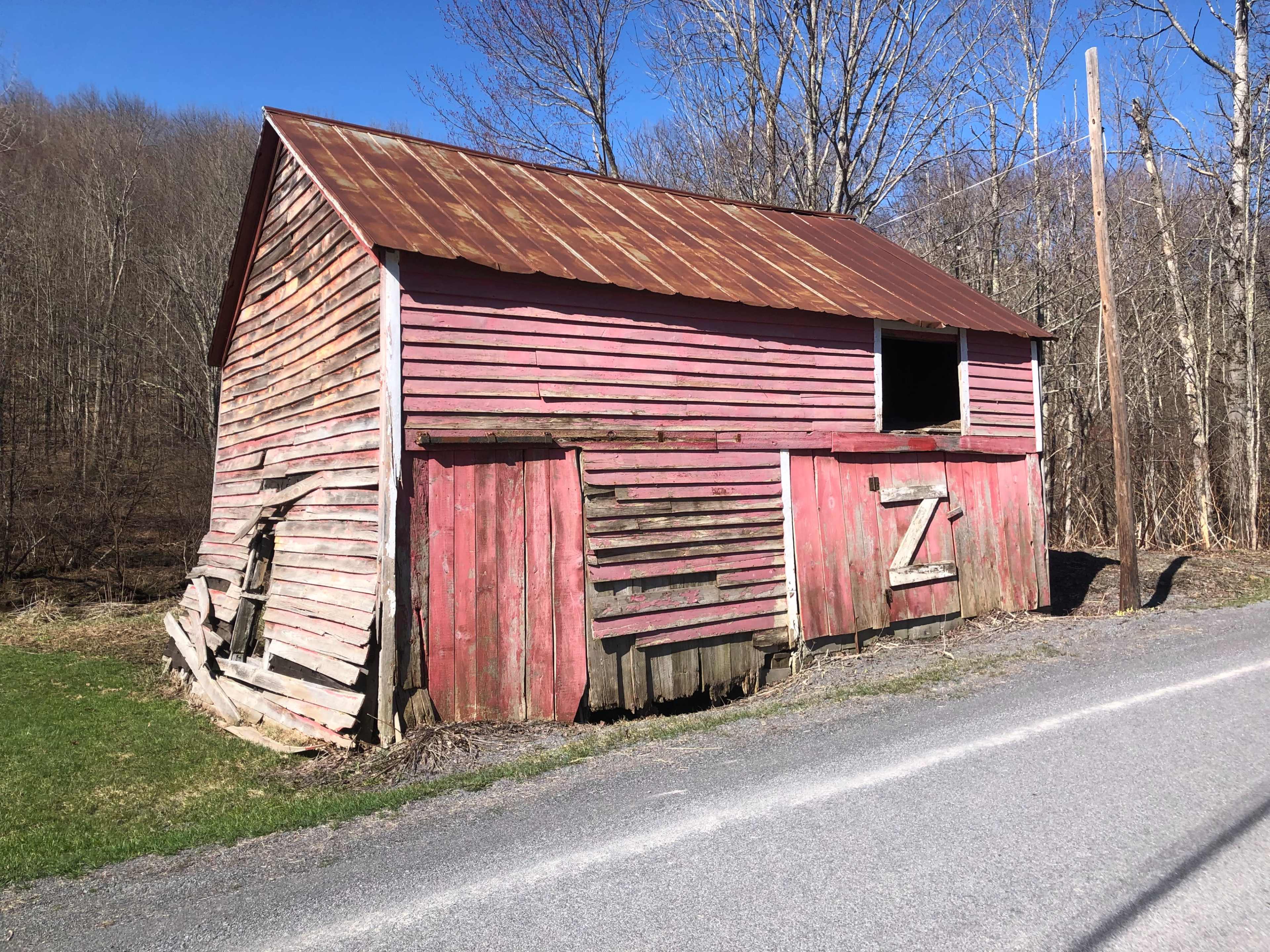 An aging, weathered red barn with a metal roof leans at an angle beside a gravel road.