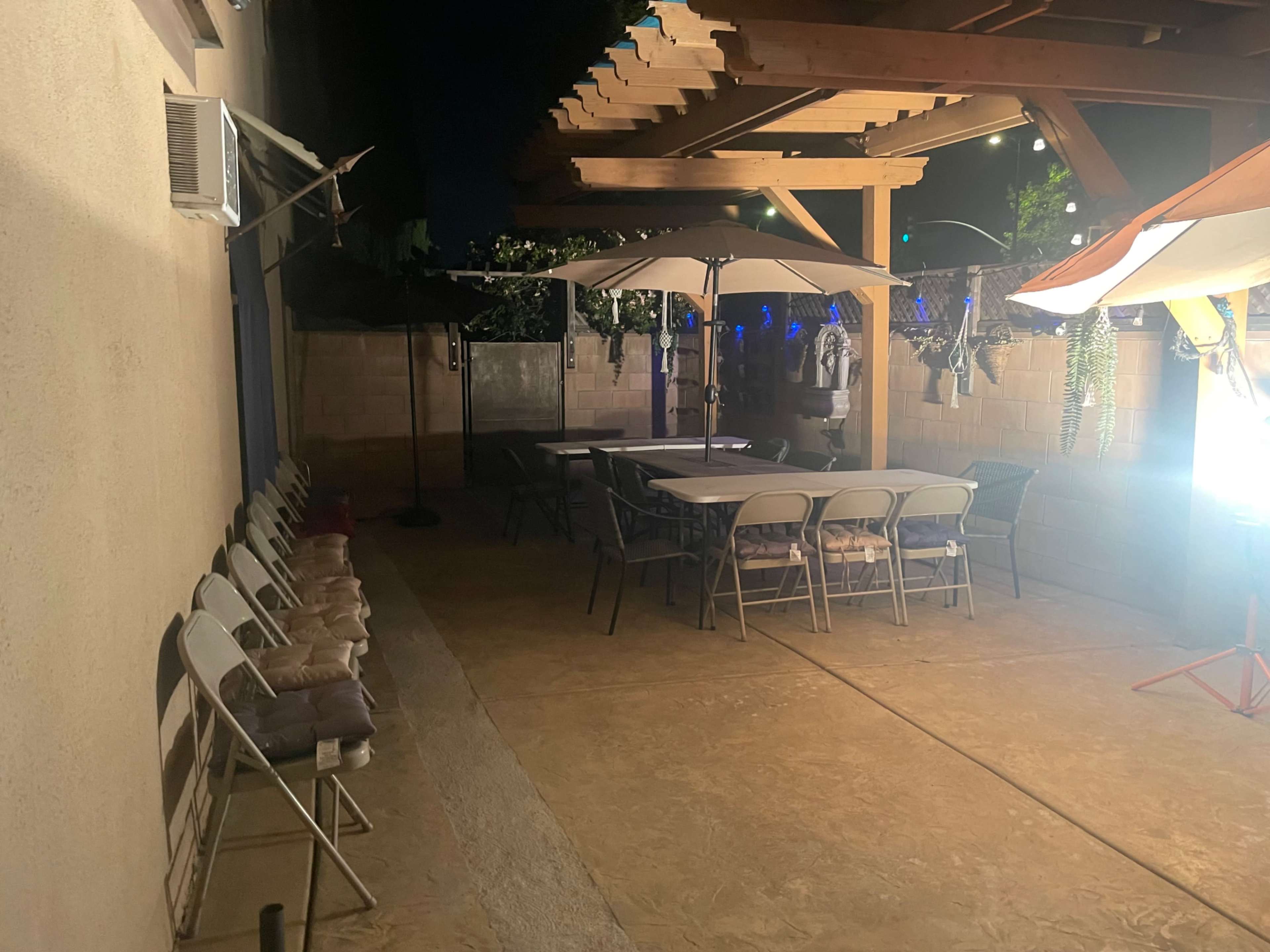 A patio area features a long table surrounded by folding chairs, with umbrellas and a pergola overhead.