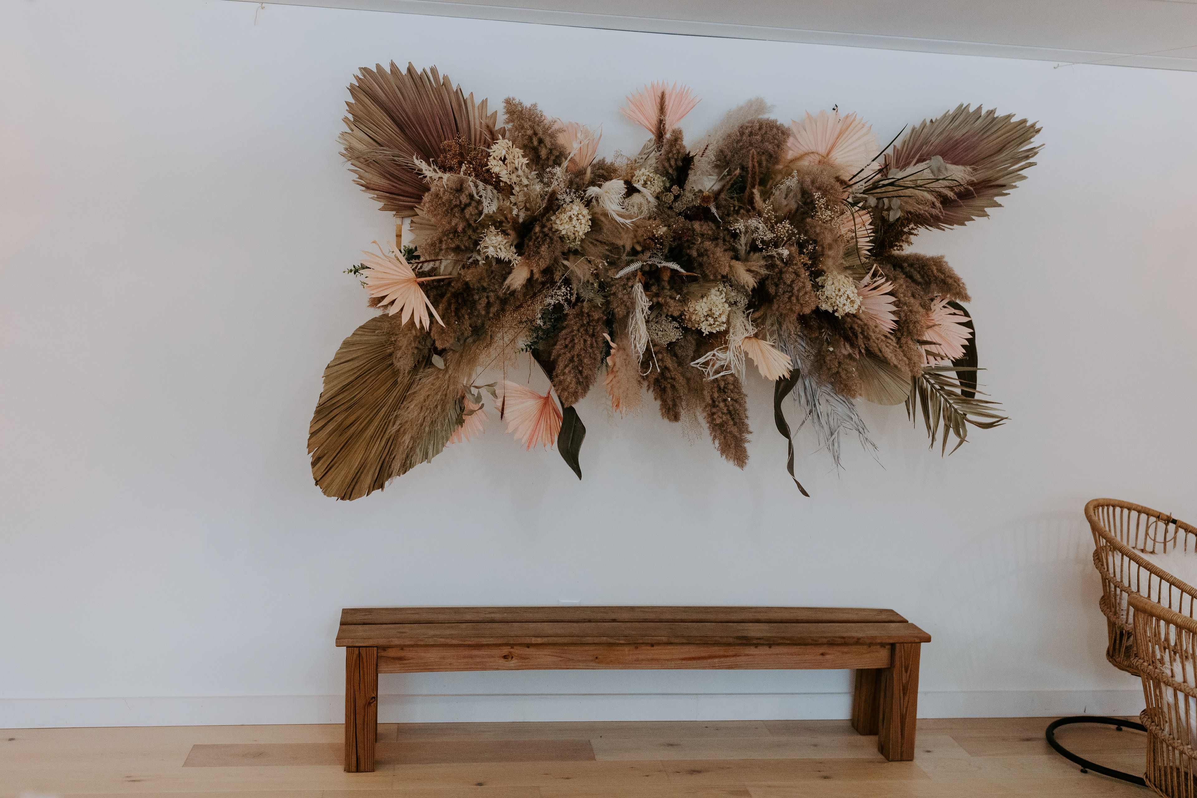 A wooden bench sits beneath a large decorative arrangement of dried plants and flowers mounted on a white wall.