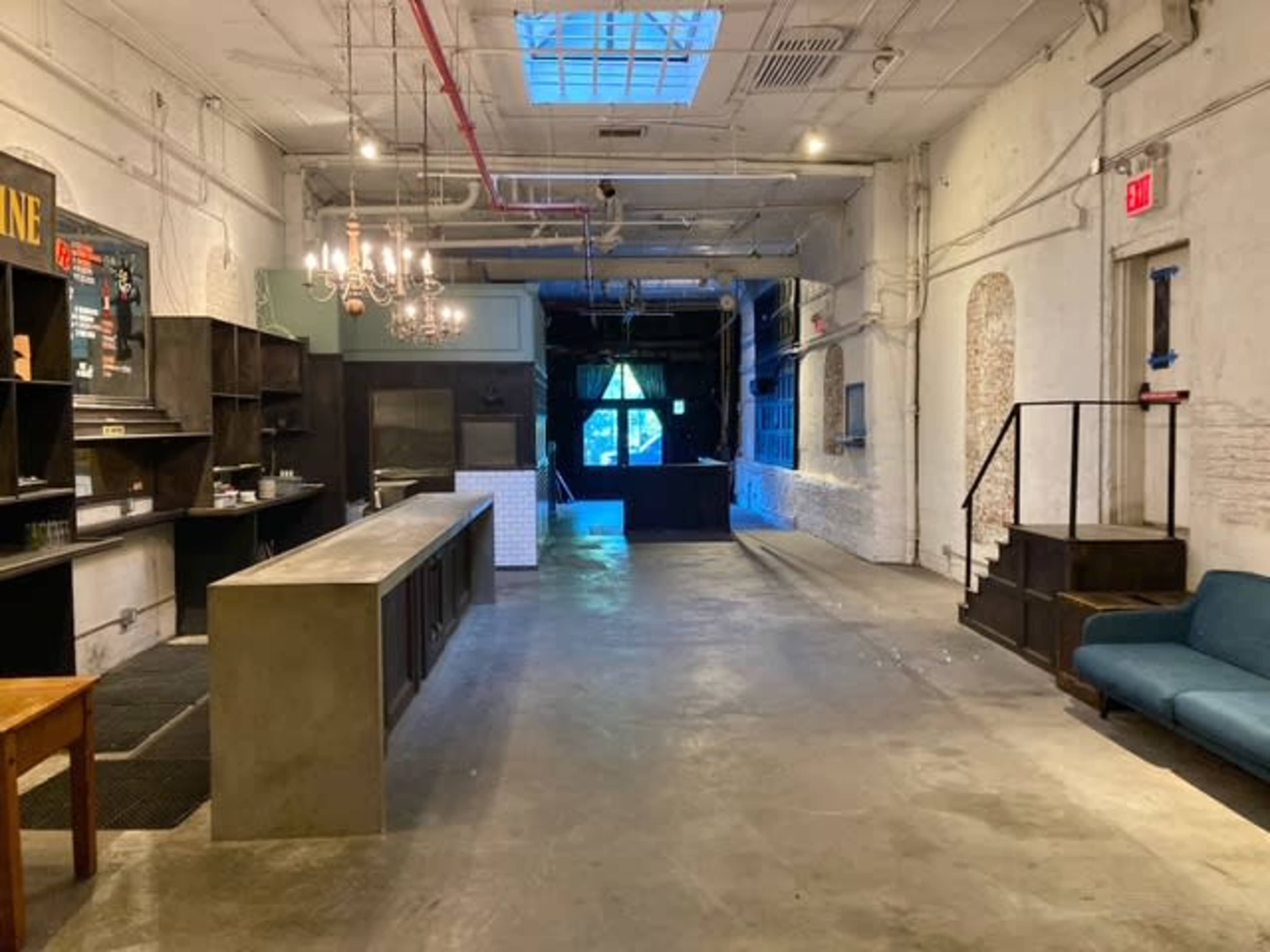 The image shows an empty, spacious interior of a modern industrial-style room with light-colored walls, a concrete bar counter, and a blue couch against a side wall.