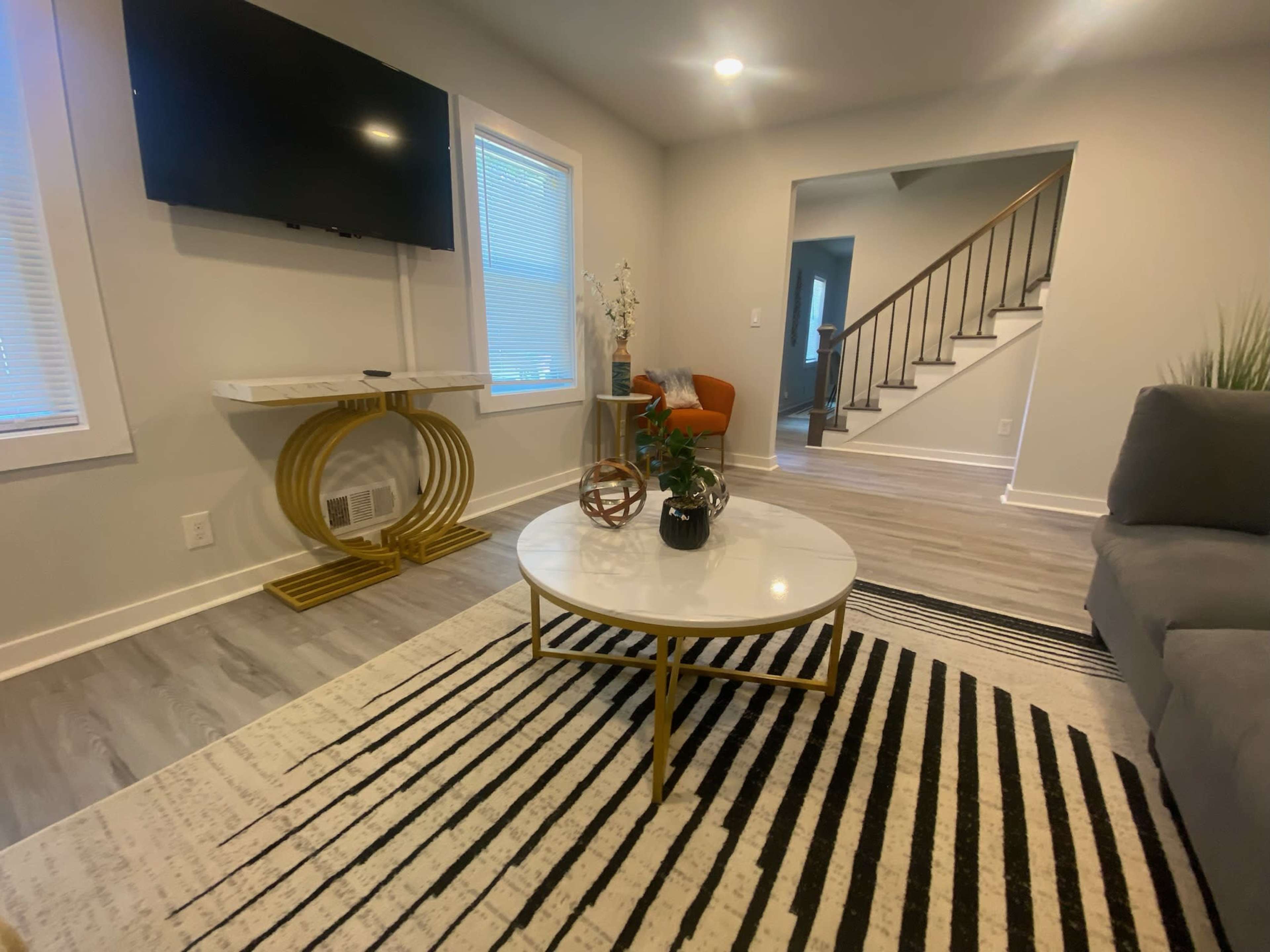 A modern living room featuring a gray sofa, a circular coffee table with a white top and gold accents, a wall-mounted television, and a decorative plant beside an orange chair.