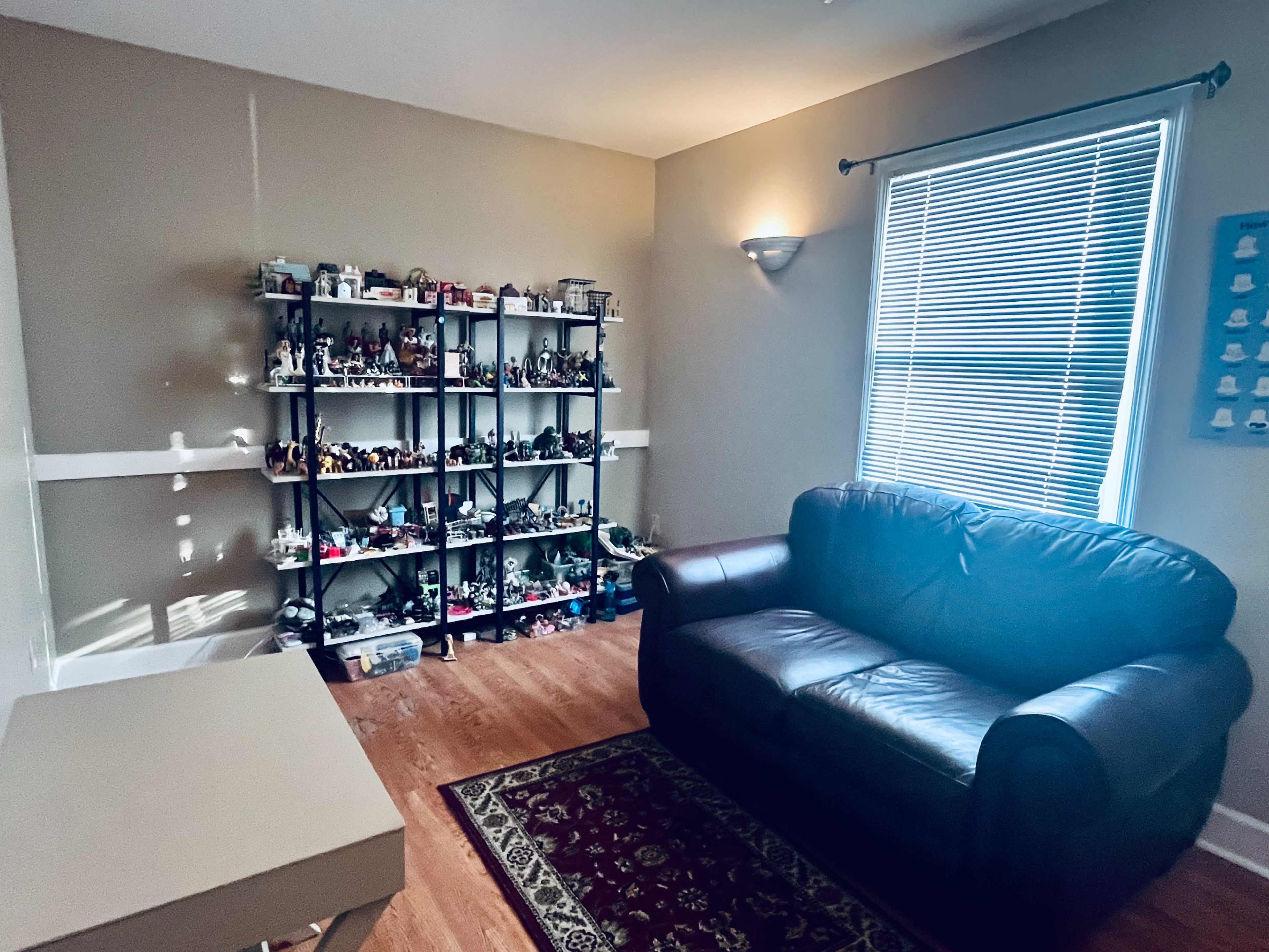 A small room features a black leather sofa, a bookshelf filled with figurines, and a window with blinds.