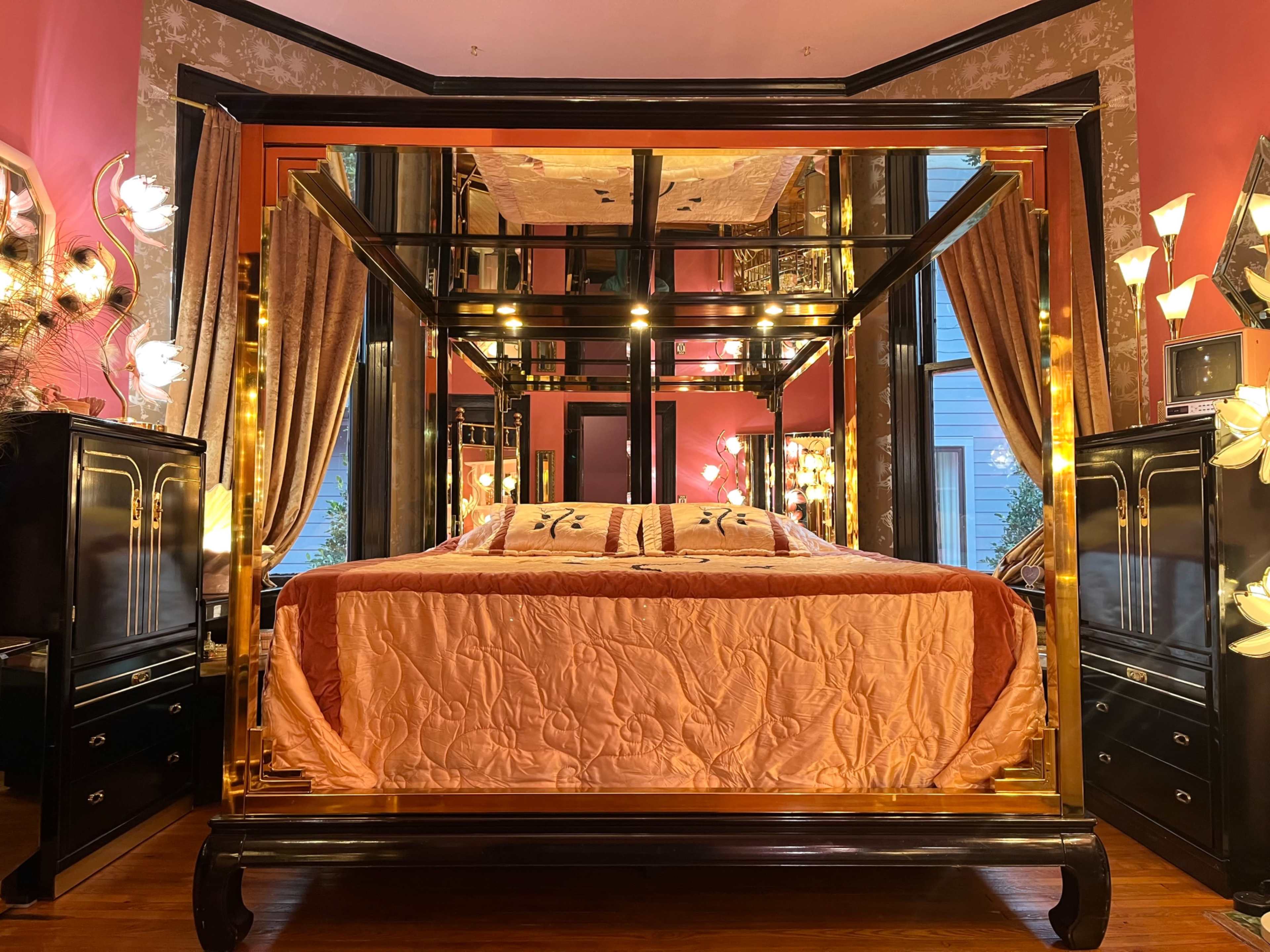 The image shows a brightly colored bedroom featuring a large, mirrored four-poster bed with an orange quilt, flanked by dark wood nightstands and warm lighting fixtures.