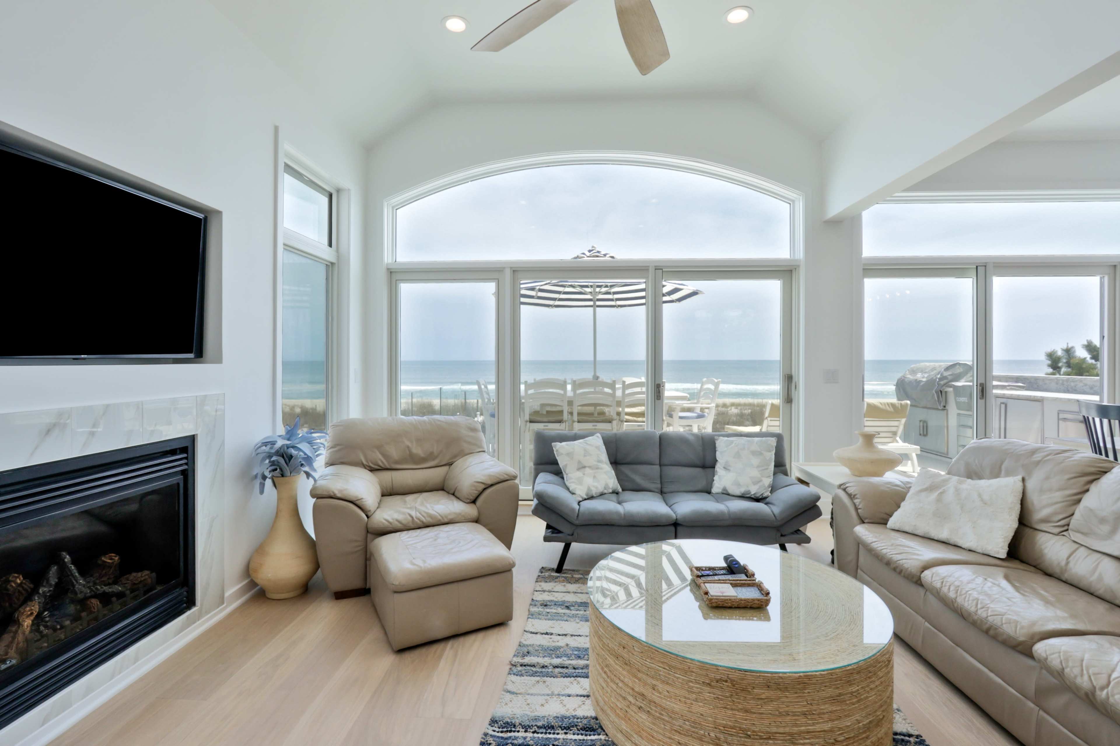 The image shows a modern living room with a view of the ocean through large windows, featuring comfortable seating and a round coffee table.