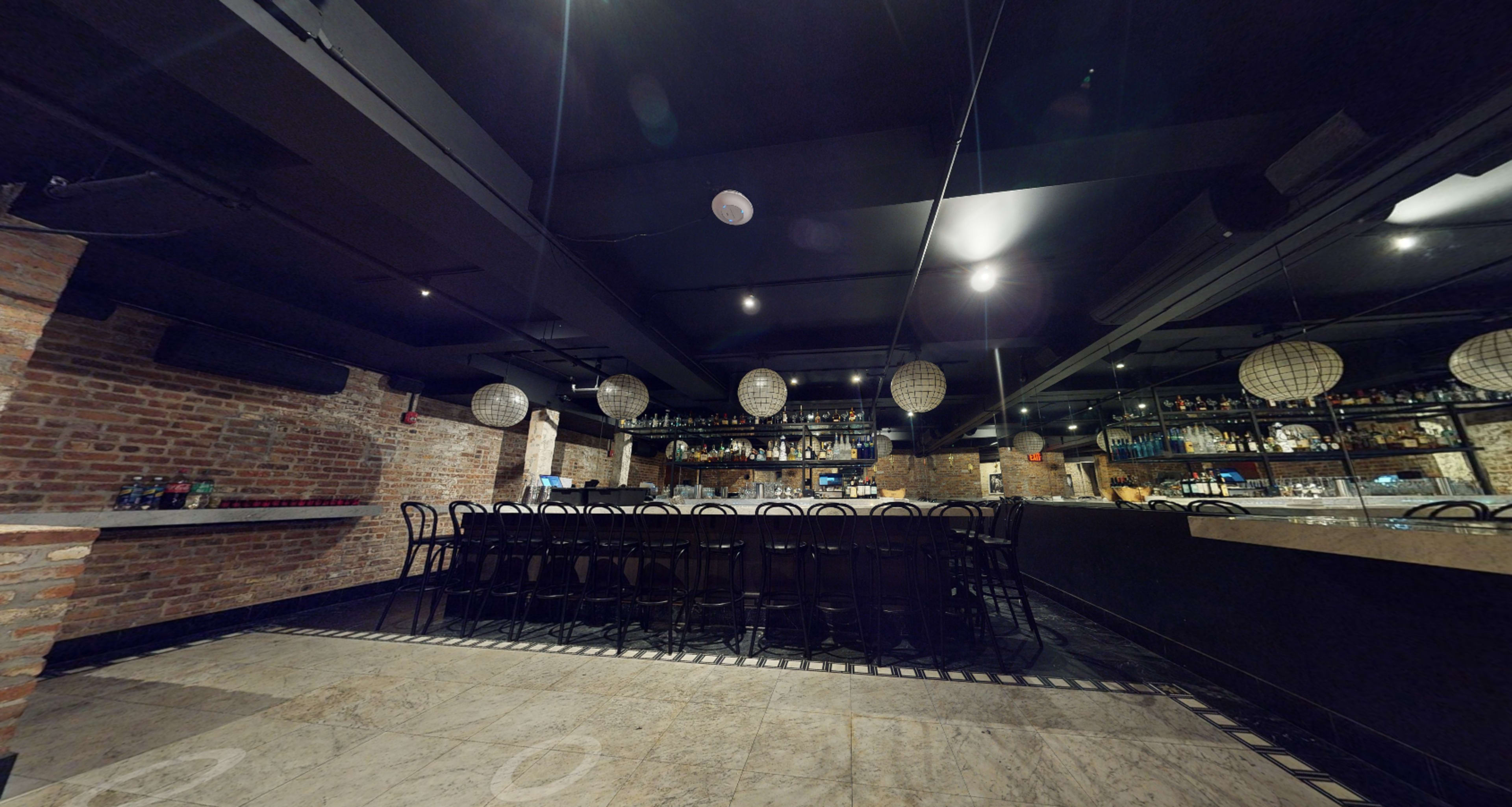 The image shows a dimly lit bar with a long seating area and exposed brick walls, featuring round hanging light fixtures and a back display of bottles.