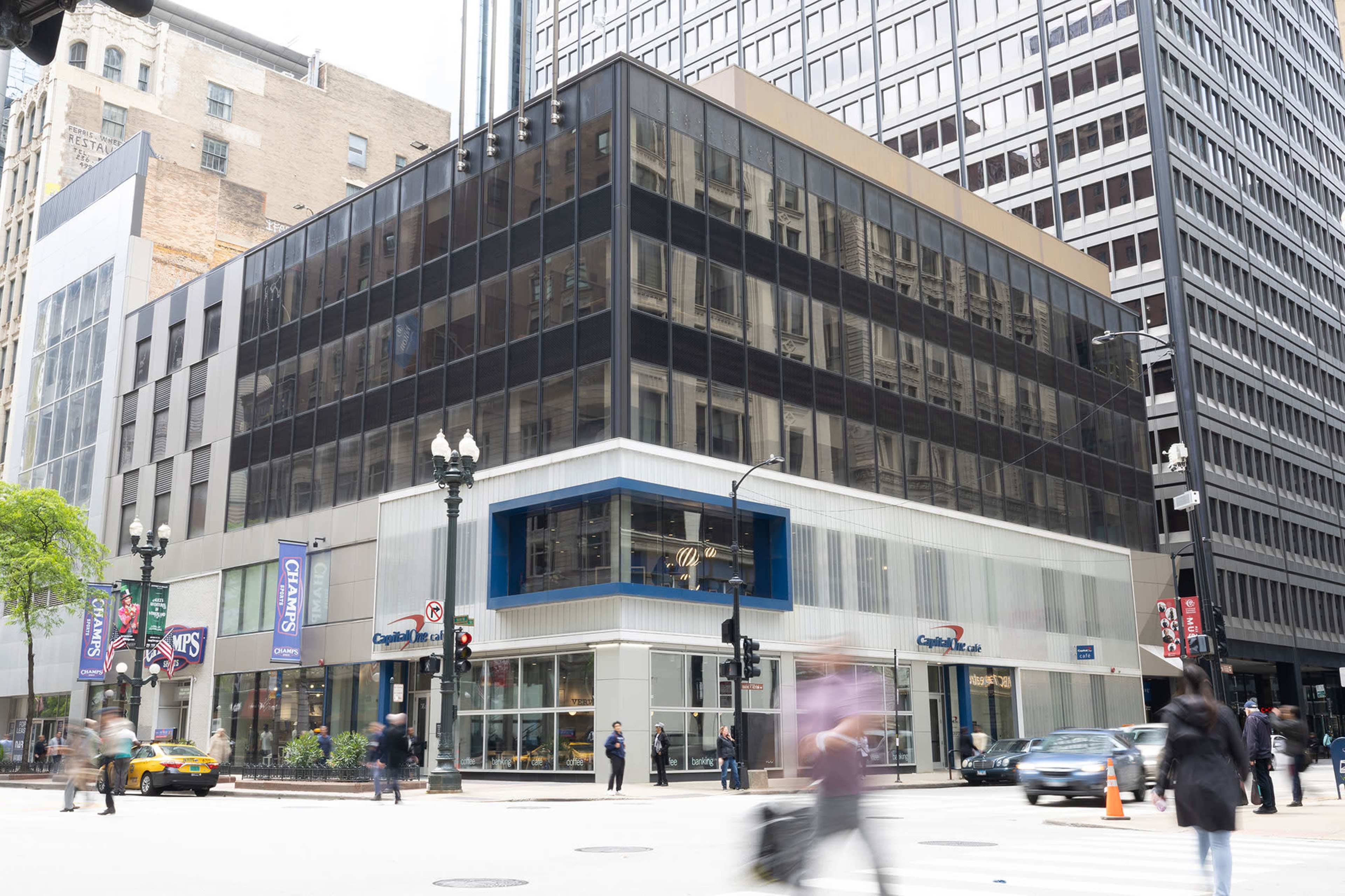 A multi-story glass building with a modern facade stands at a busy street corner, featuring retail spaces on the ground floor and pedestrians crossing the street.