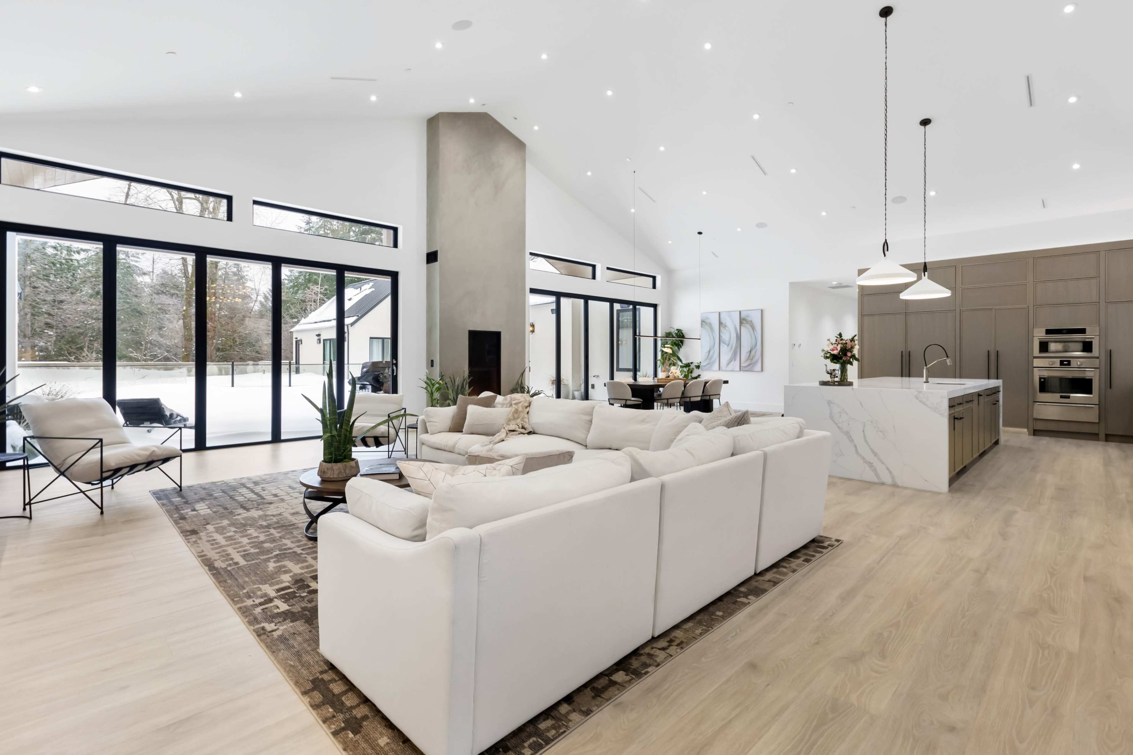 The image depicts a spacious, modern living area featuring a large white sectional sofa, a marble kitchen island, and expansive windows that provide a view of the outdoors.