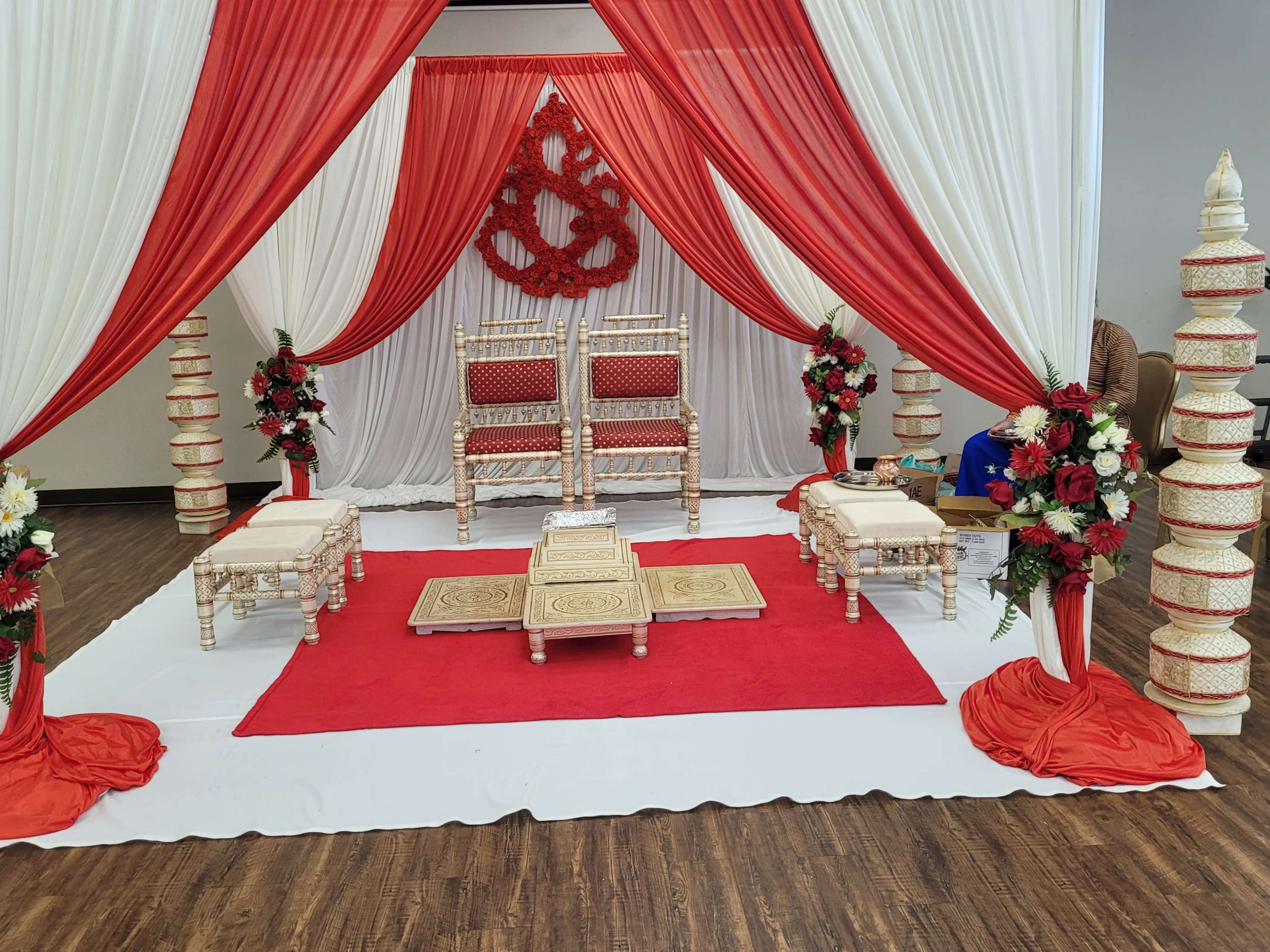 A decorated event space featuring red and white drapery, a seating area with ornate chairs, and floral arrangements on either side.