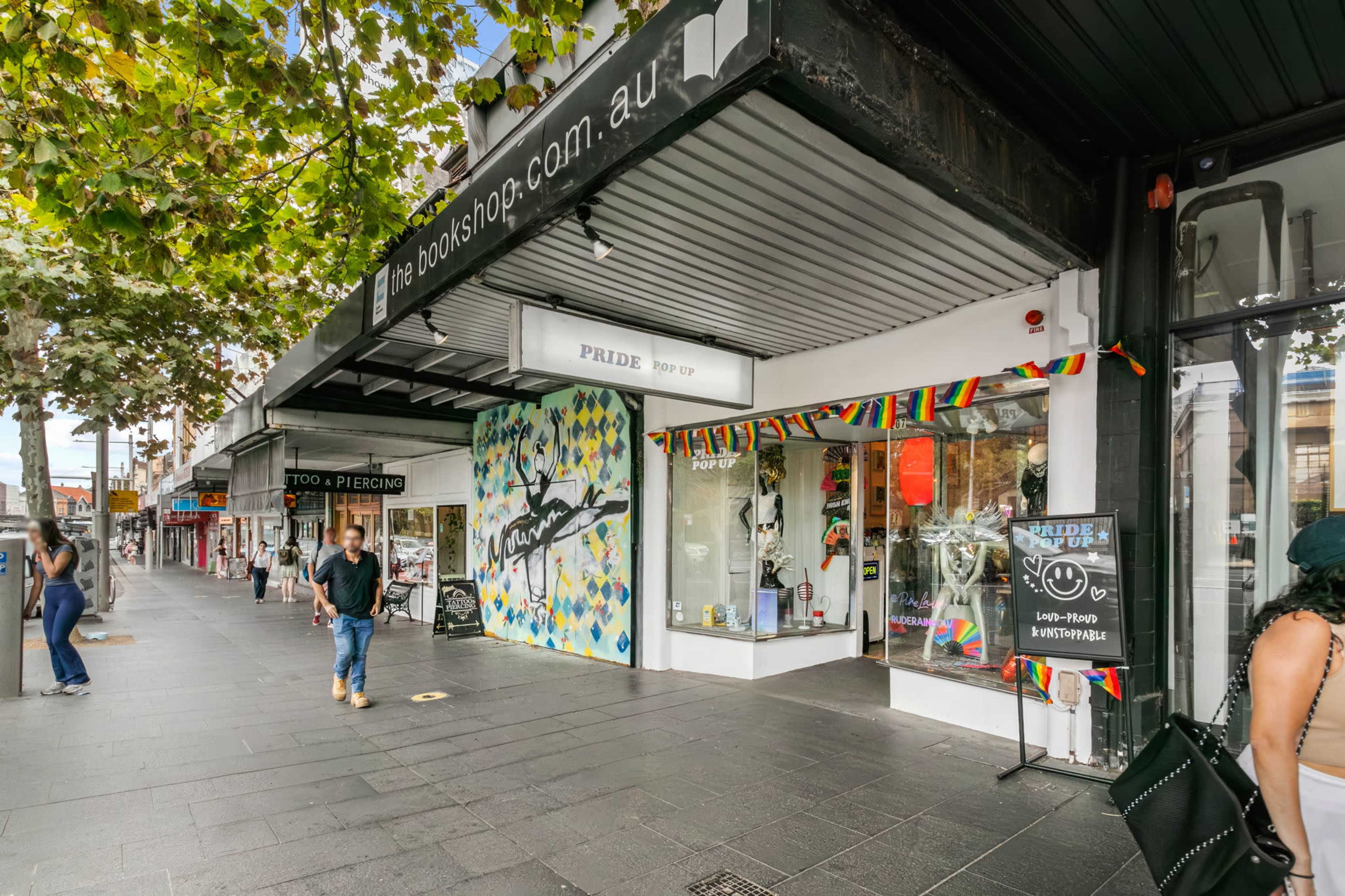 A shopfront displays colorful decorations and a rainbow banner, with pedestrians walking on a tree-lined sidewalk.