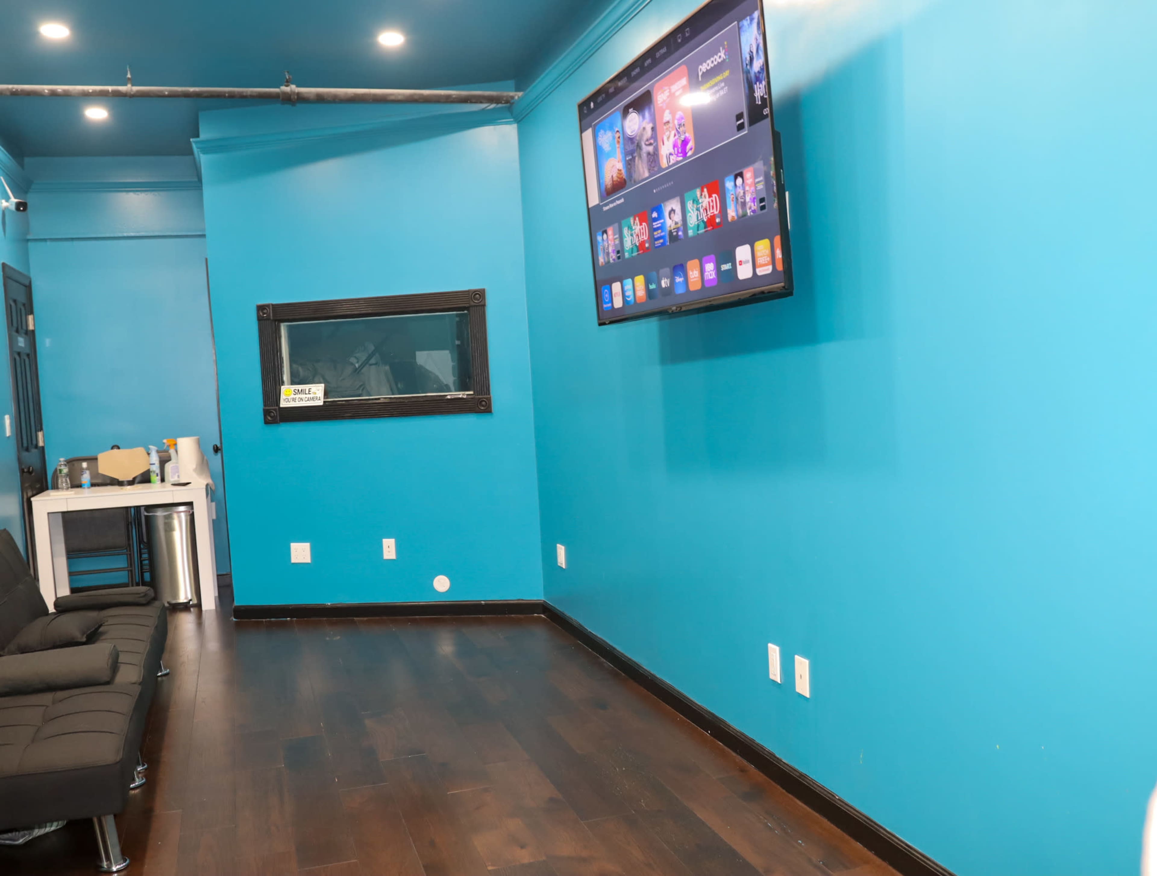 A bright blue room with a wall-mounted television displaying apps, a black sofa on the left, and an observation window on the right.
