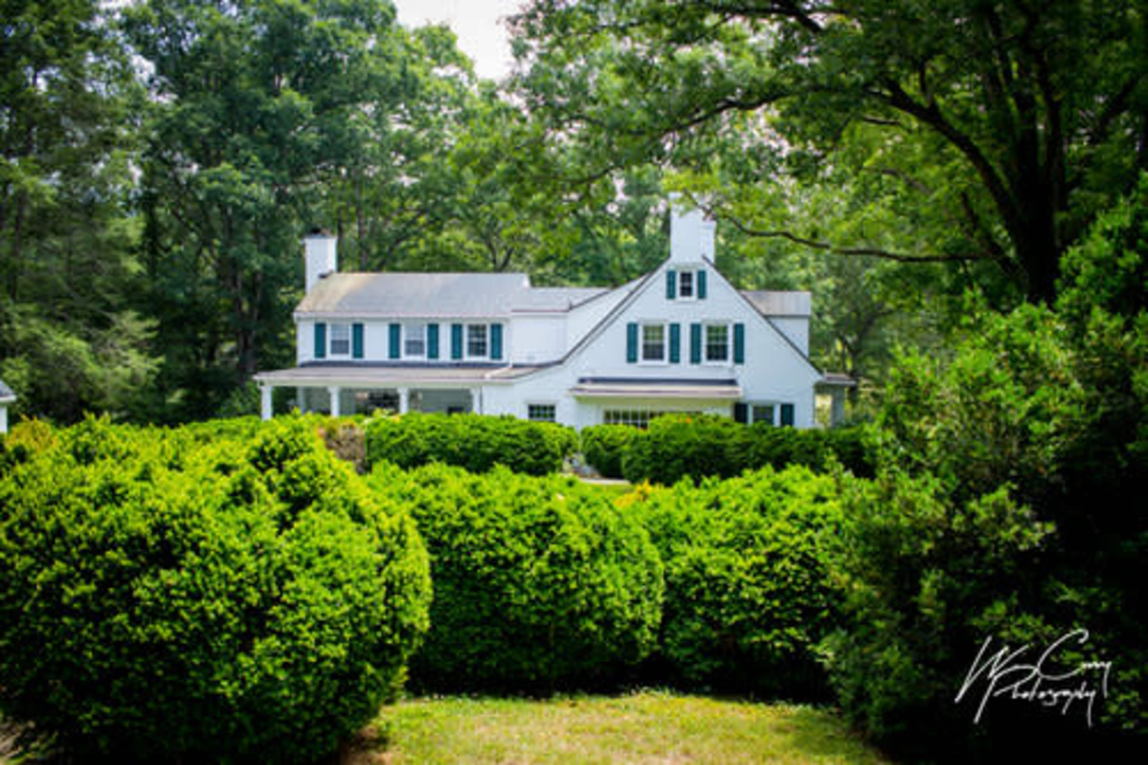 A white two-story house with green shutters is surrounded by neatly trimmed hedges and trees.