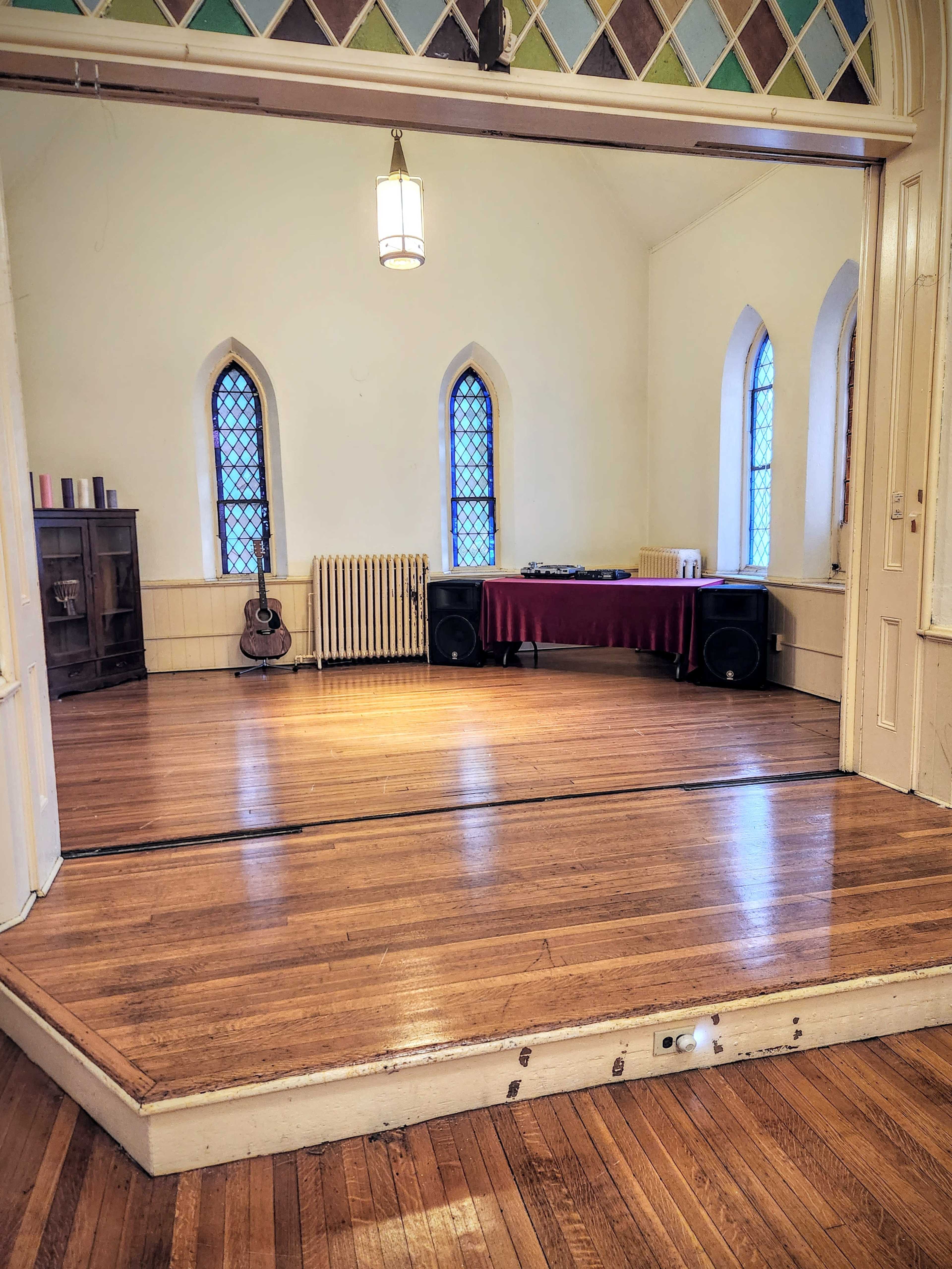 The image shows a spacious room with wooden floors, a long table covered with a red tablecloth, and guitars leaning against the wall near windows with decorative glass.