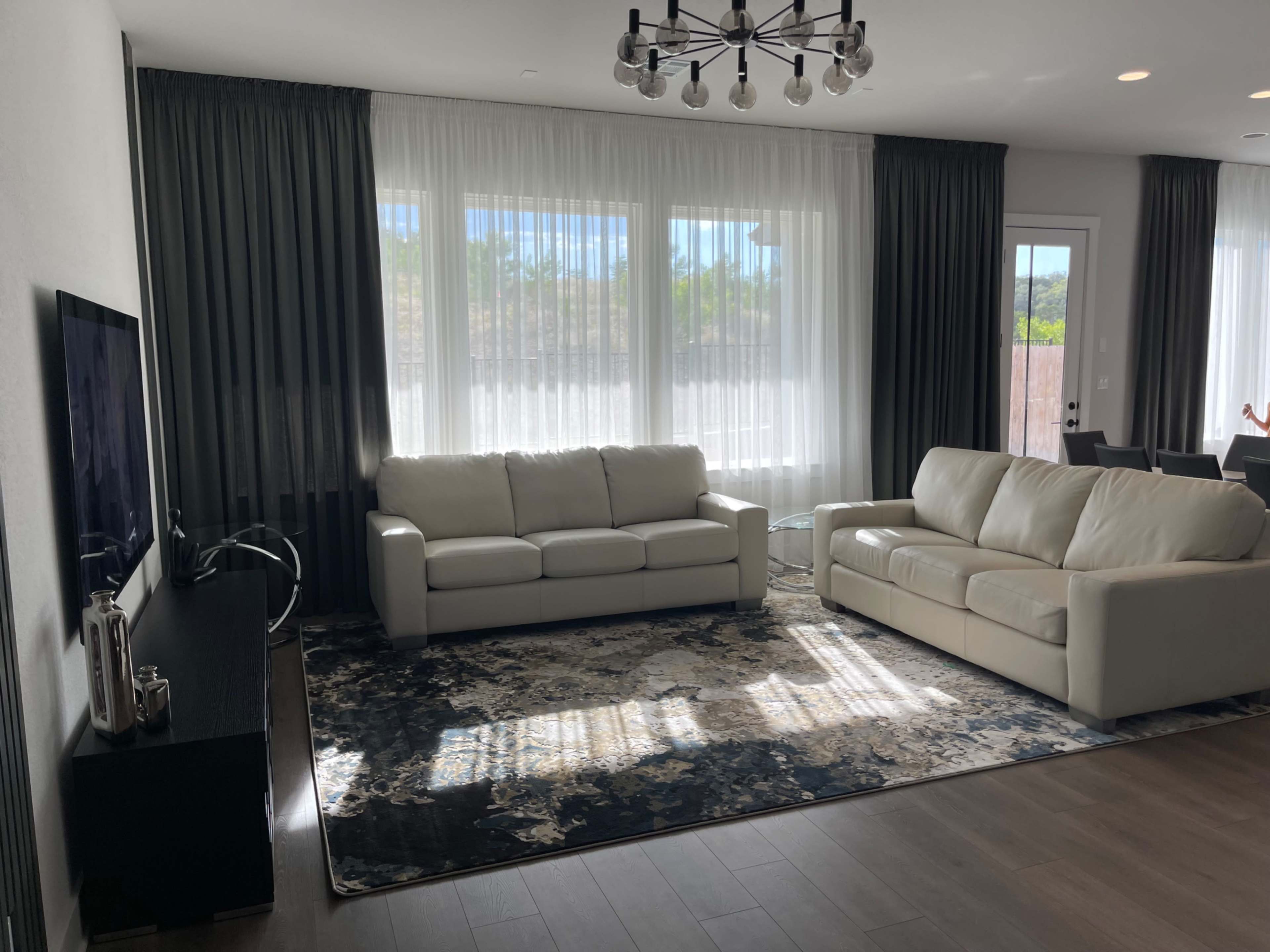 The living room features two white sofas, a dark area rug, a television on a black cabinet, and large windows with sheer curtains.