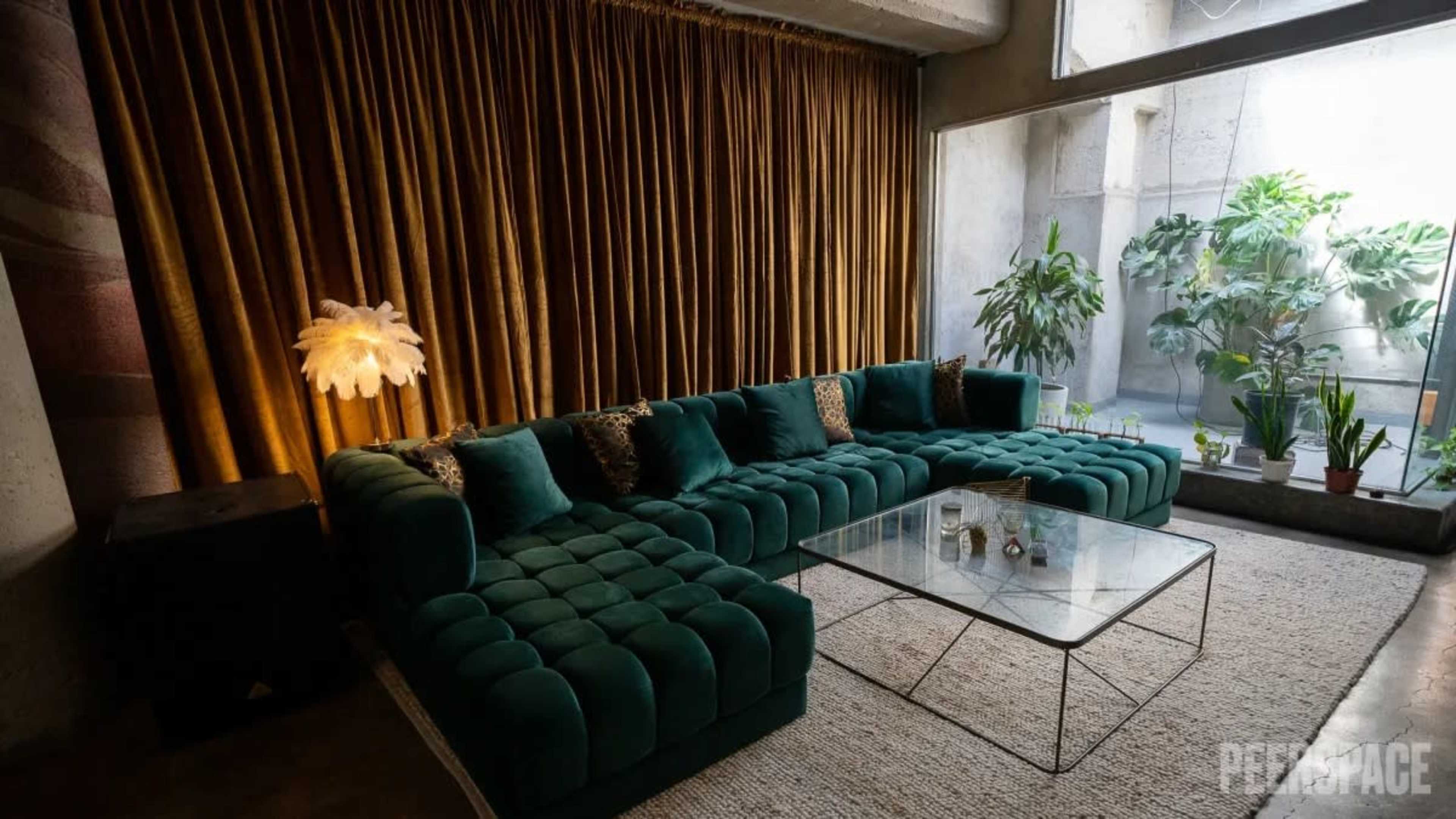A modern living room with a large teal sectional sofa, a glass coffee table, and potted plants near a window draped with brown curtains.