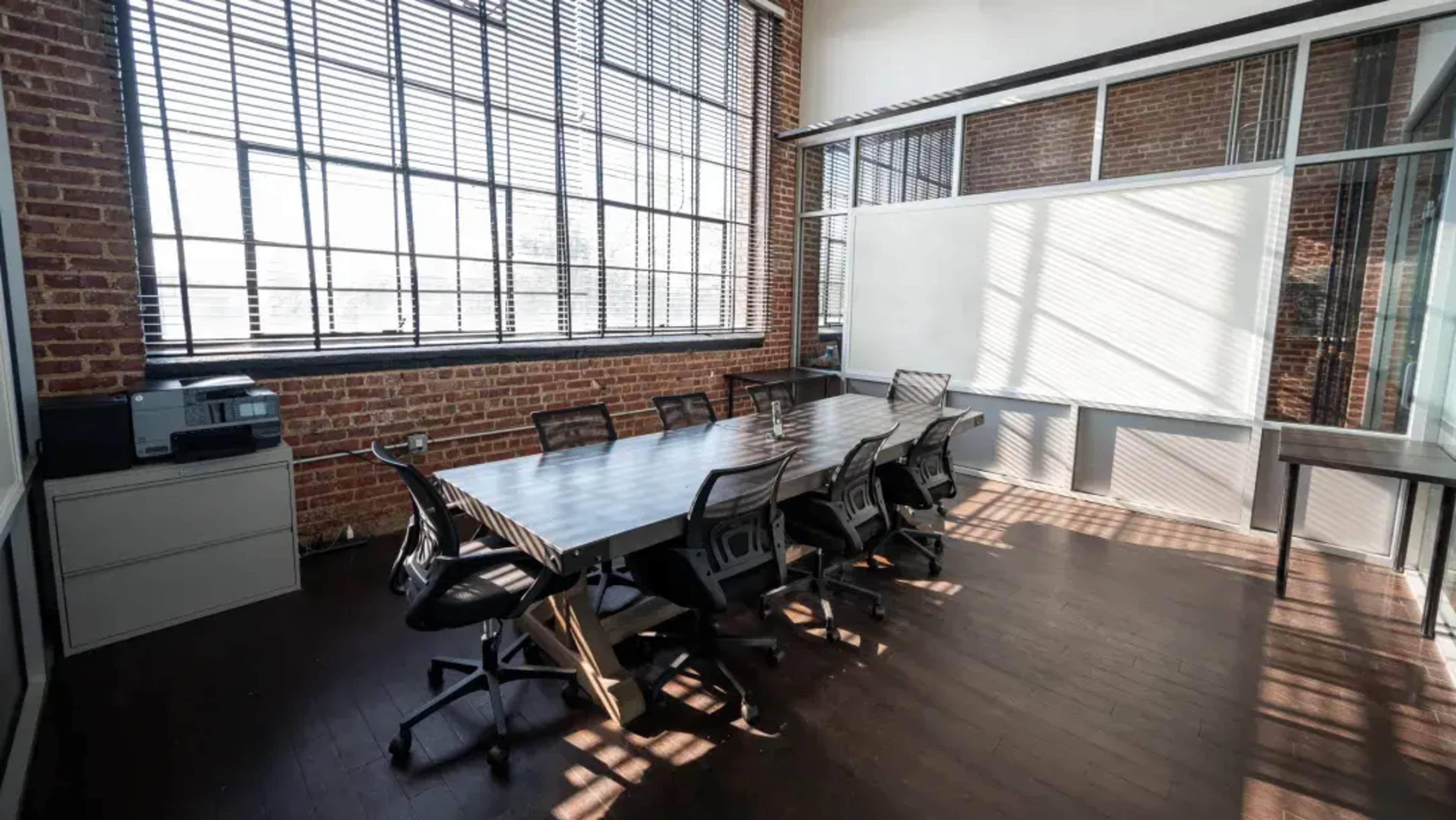 The image shows a modern conference room featuring a long table surrounded by several mesh office chairs, with large windows allowing natural light to fill the space.