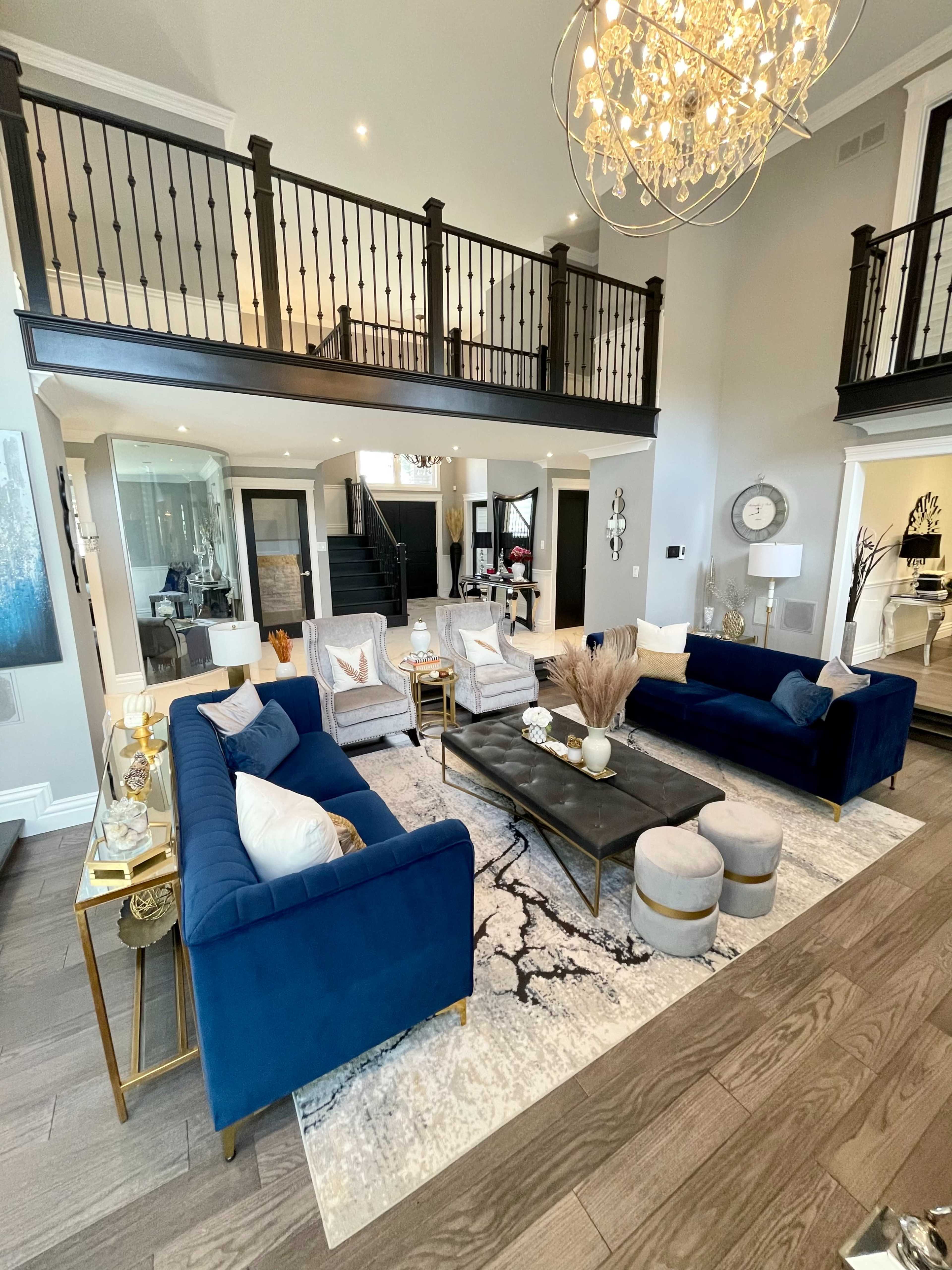 A spacious living room with blue sofas arranged around a black coffee table, an area rug, and a chandelier overhead, complemented by a stylish staircase in the background.