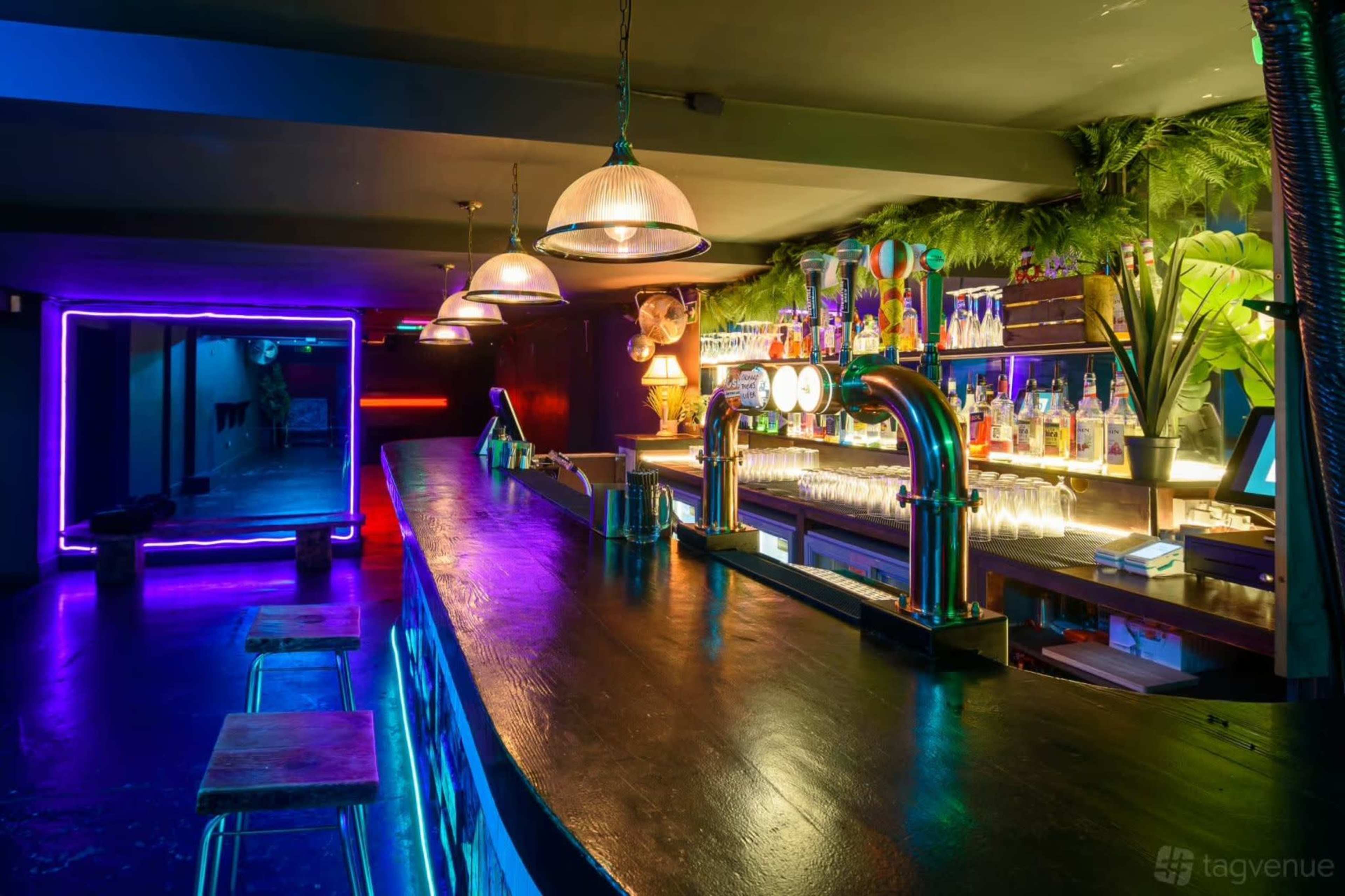 The image shows a modern bar with bright neon lights, a long wooden counter, and a variety of colorful drinks displayed behind it.