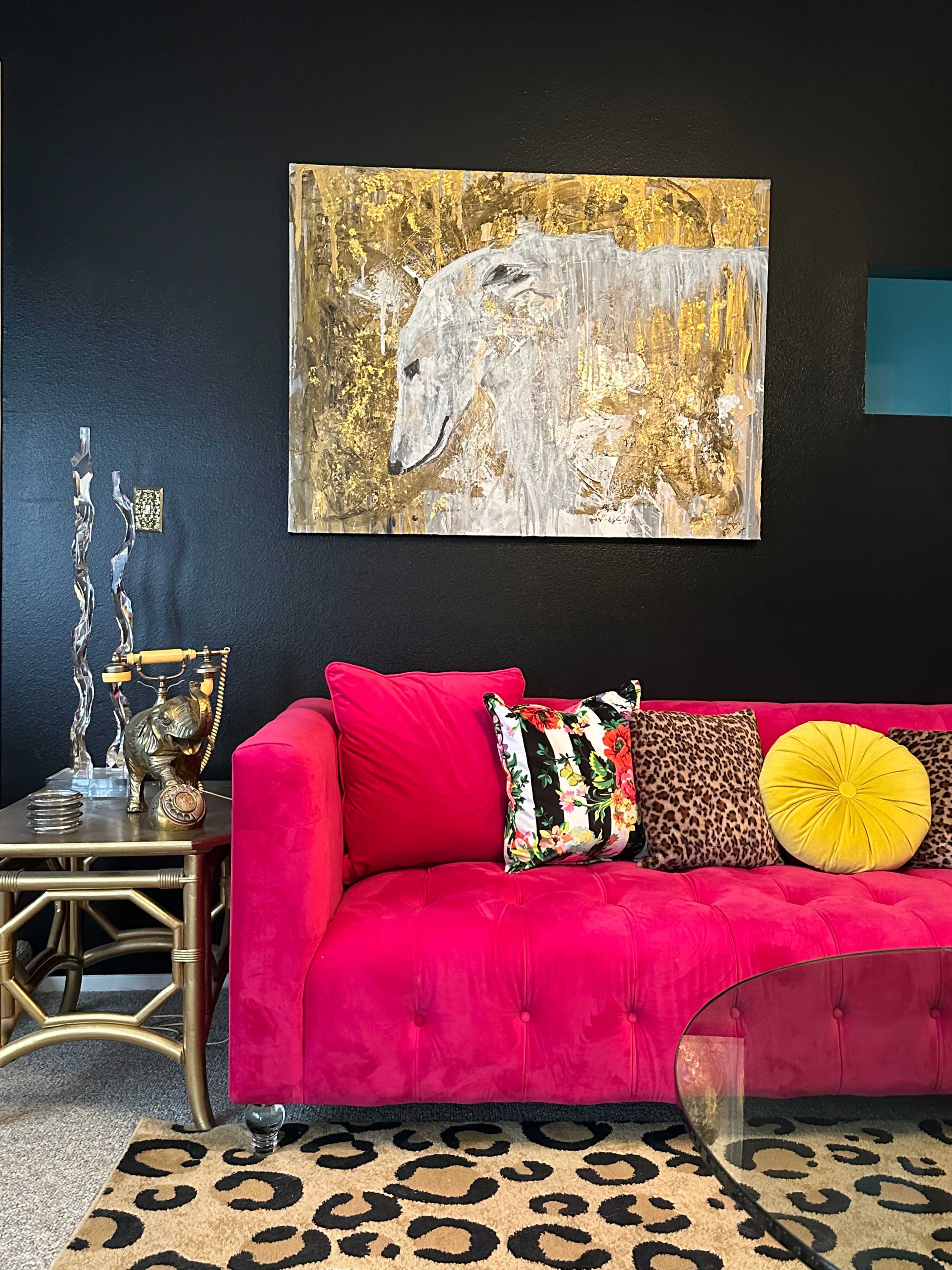 A bright pink couch adorned with patterned pillows, positioned against a black wall, with a gold and gray painting of a dog above it, accompanied by a glass coffee table and decorative elements nearby.