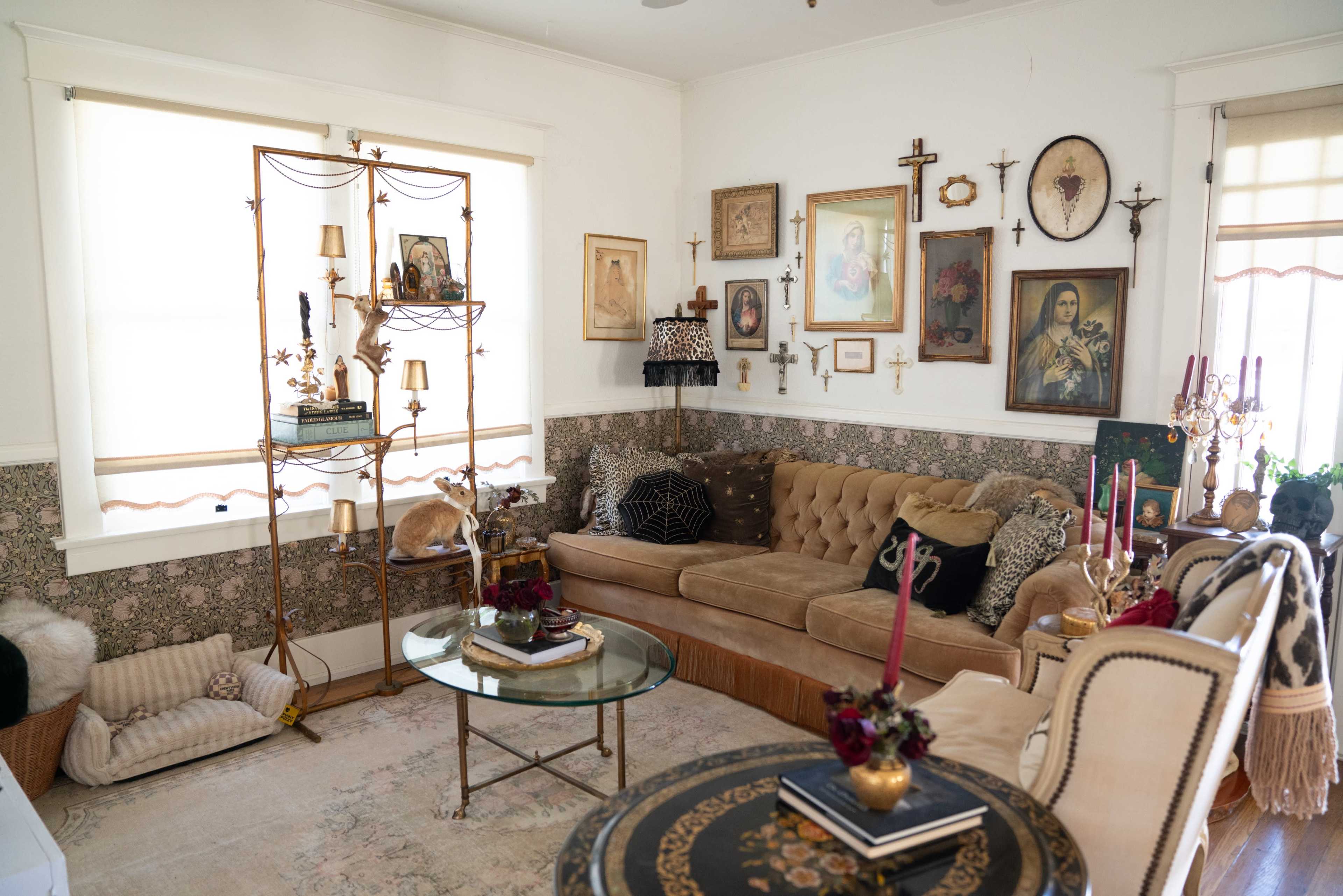 A cozy living room features a brown velvet sofa, various framed artworks on the walls, a glass coffee table, and decorative candles arranged on a side table.
