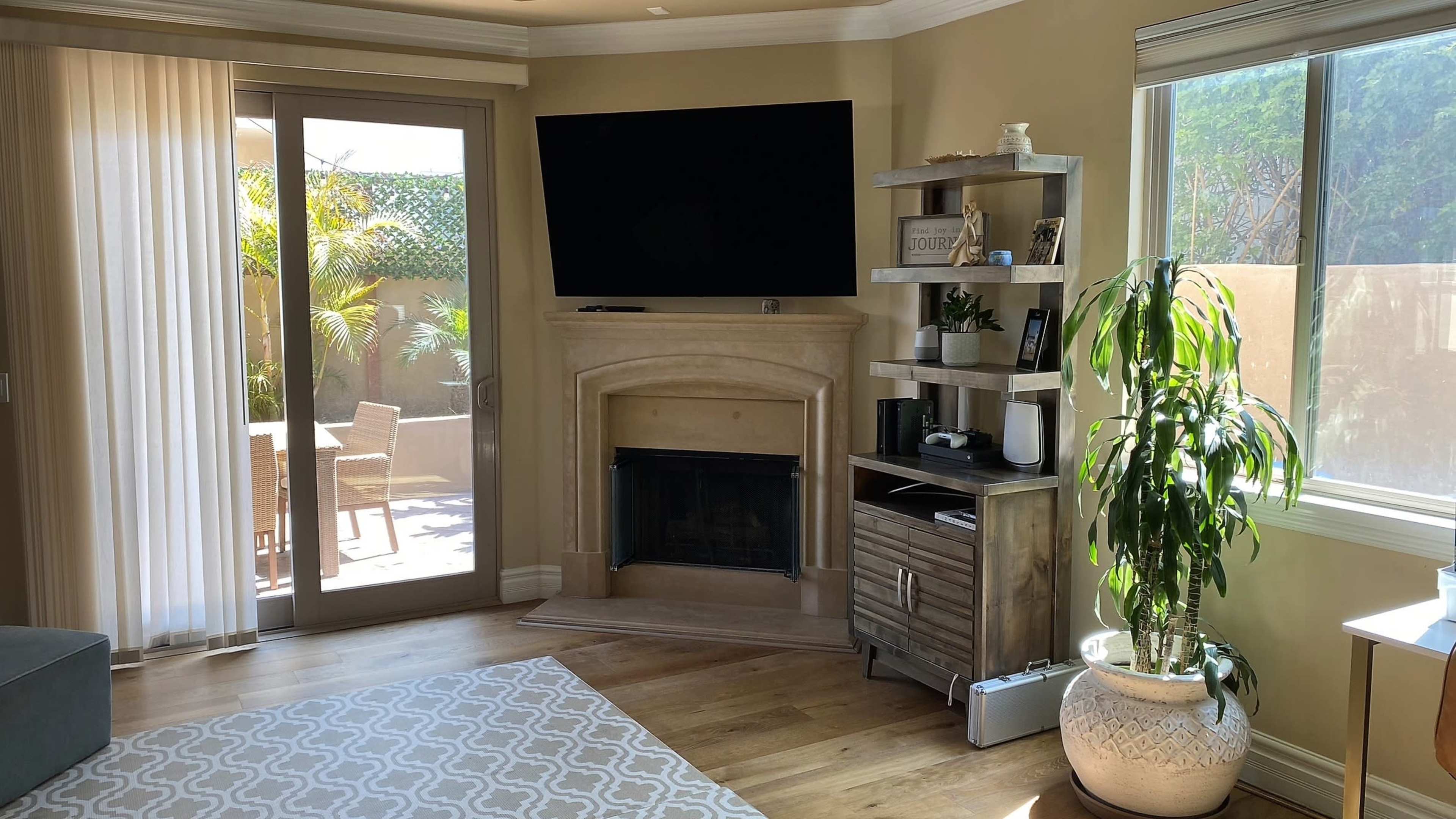 A living room features a television mounted above a fireplace, with a large window revealing an outdoor patio and a potted plant in the corner.