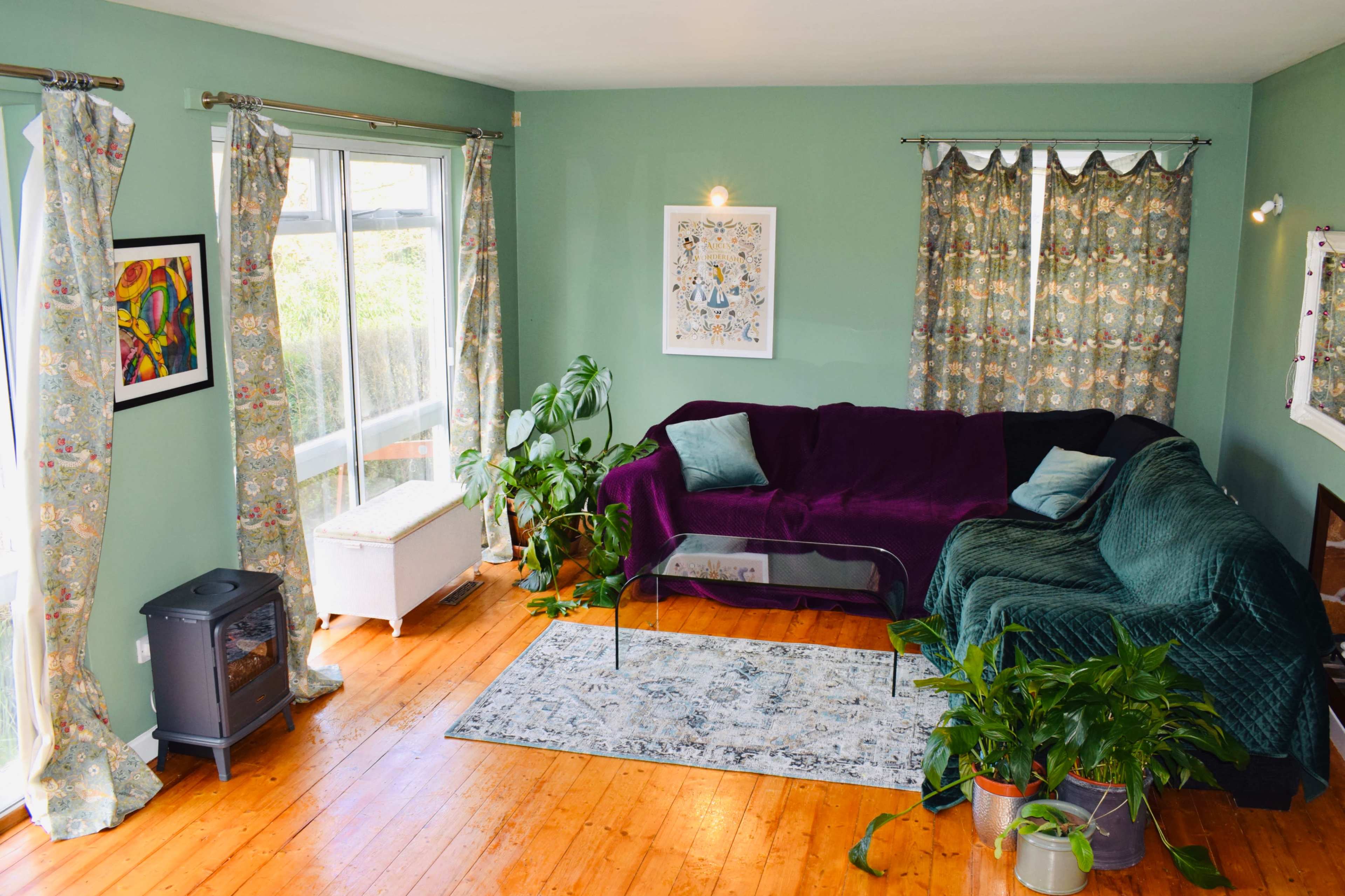 The living room features two couches, a coffee table, a rug, several plants, and large windows with decorative curtains.