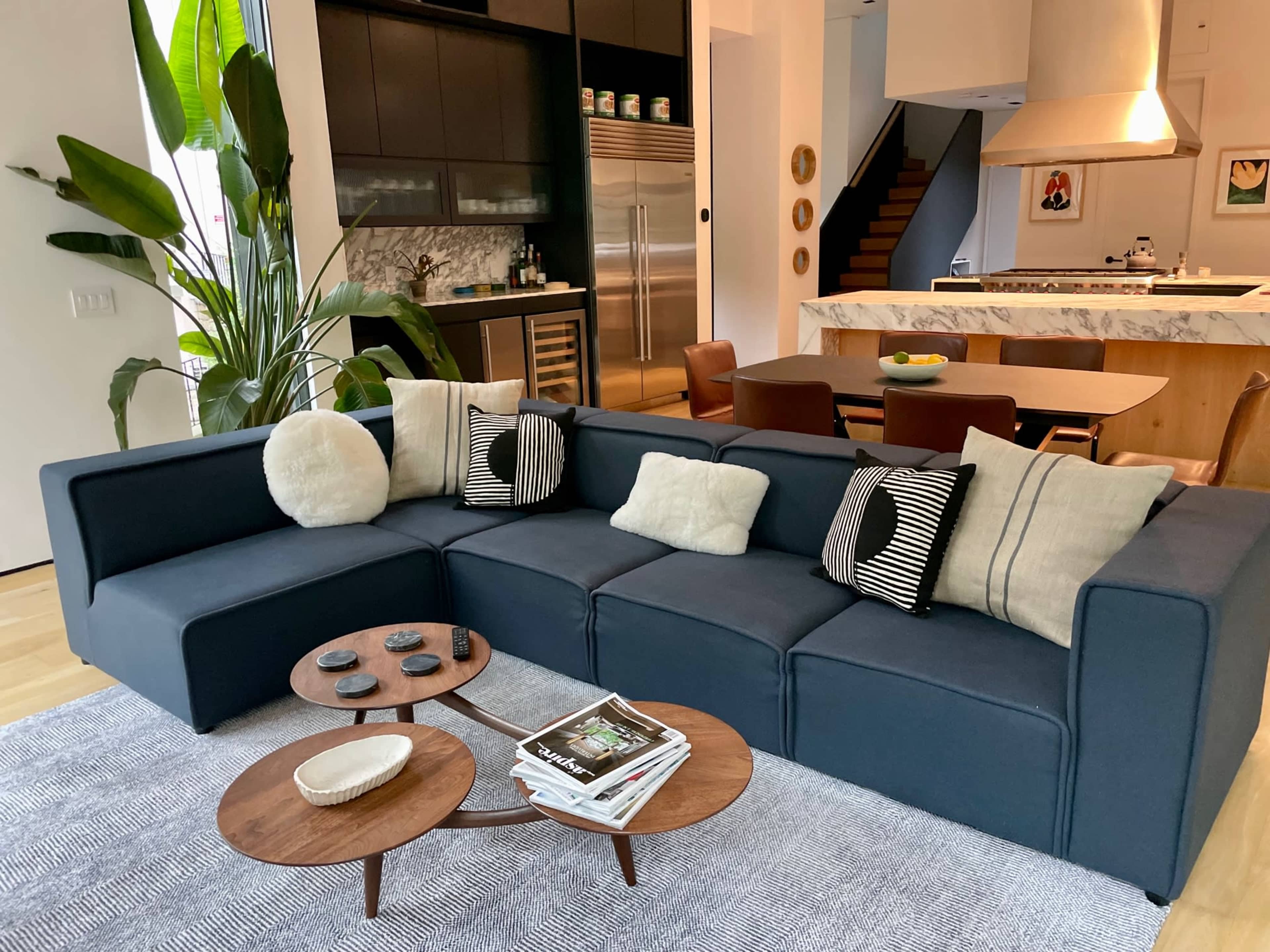 A modern living room features a dark sectional sofa with patterned pillows, a pair of wooden coffee tables, and a kitchen visible in the background.