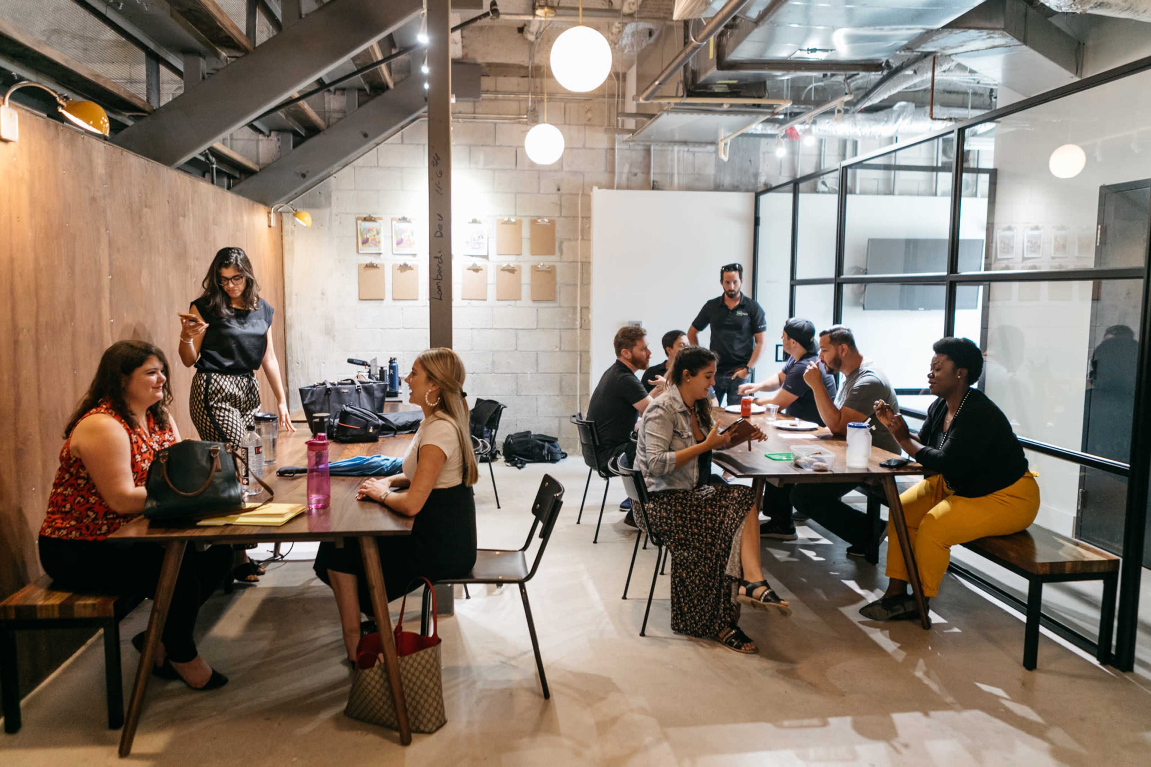 A group of people are engaged in discussions at various tables in a modern office space with industrial elements.