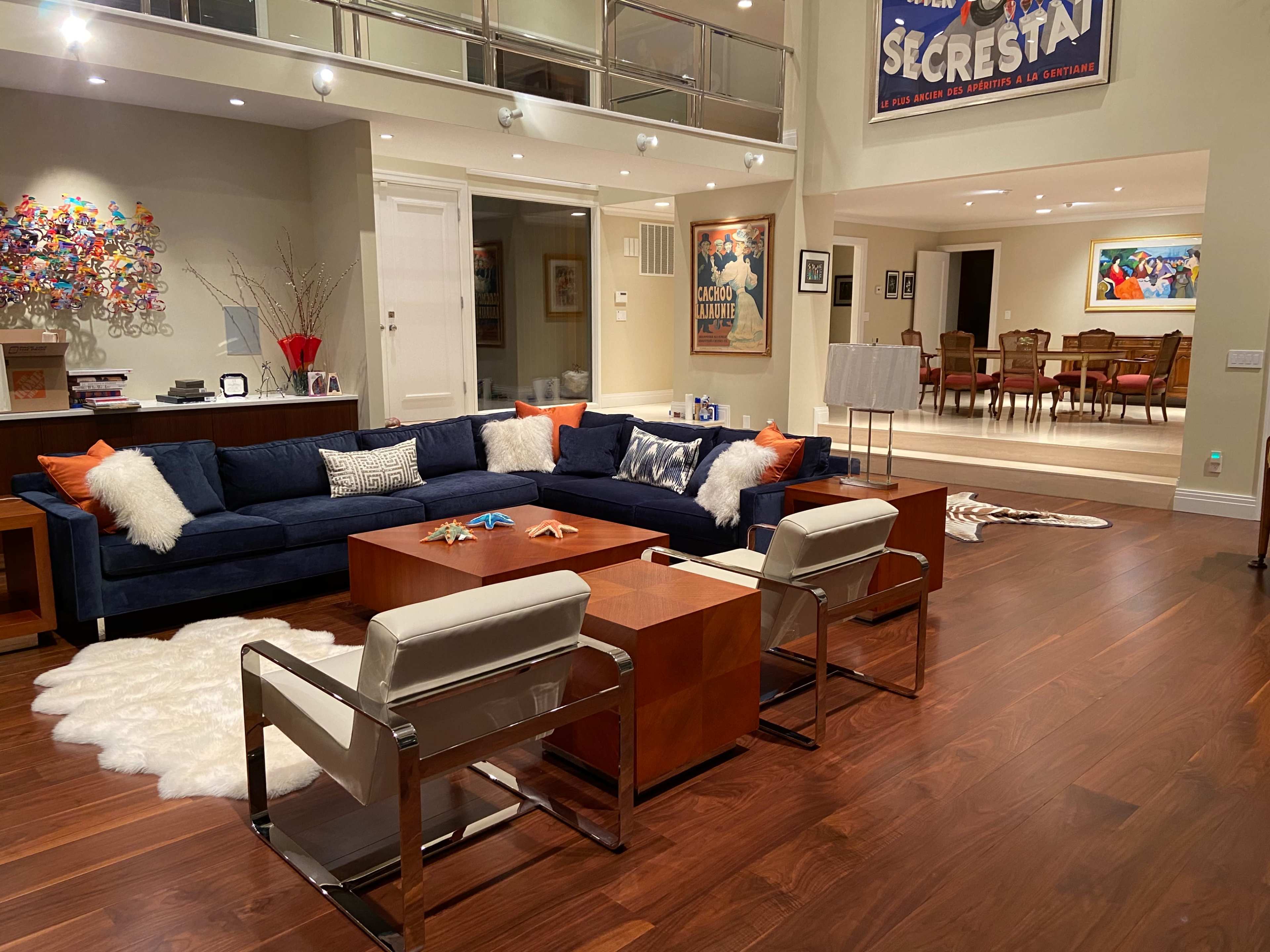 The image shows a modern living room with a large blue sectional couch, wooden coffee tables, and an arrangement of art and furniture in a spacious area with hardwood flooring.