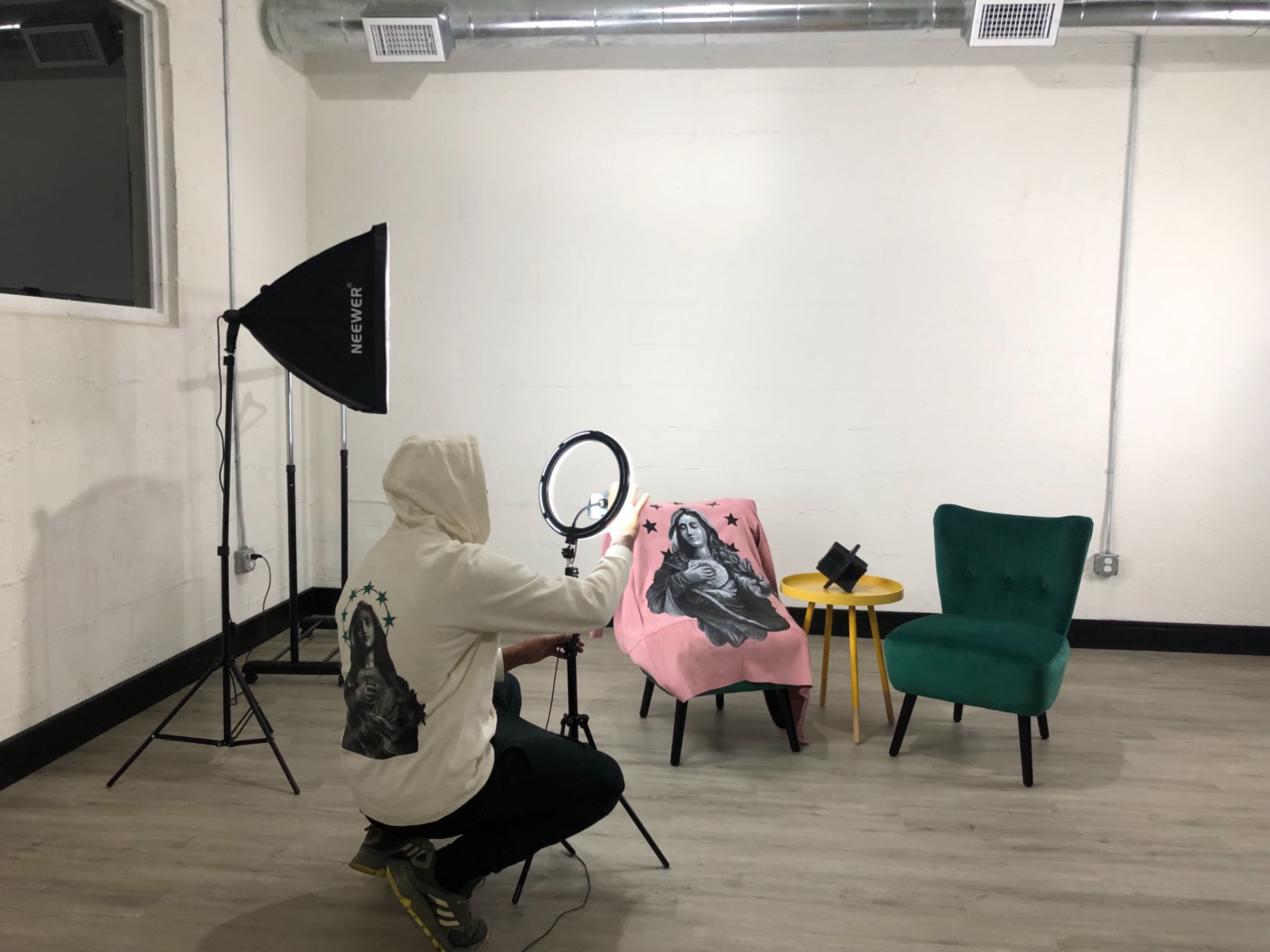 A person in a hoodie adjusts a ring light in a photography studio featuring two chairs and a pink blanket with a graphic design.