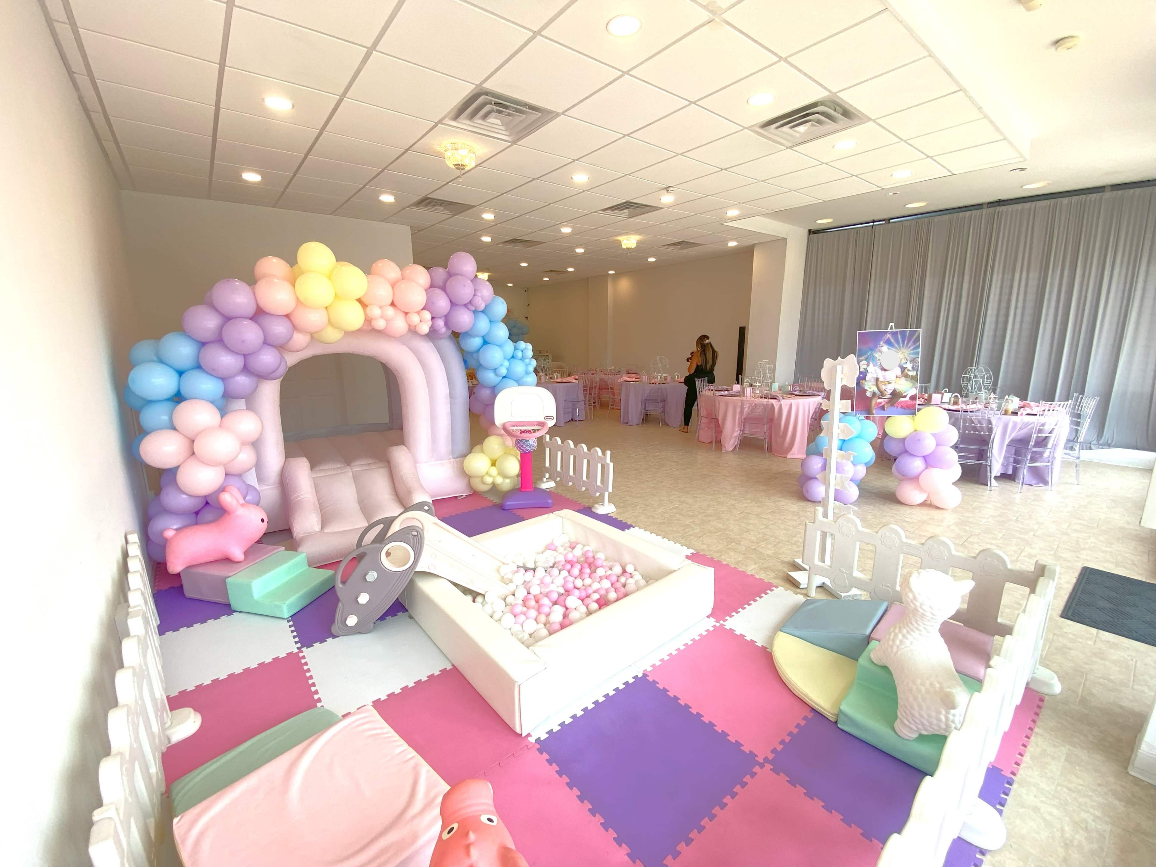 The image shows a brightly decorated children's play area filled with colorful balloons, a ball pit, and play structures, alongside tables set for a party in a softly lit room.