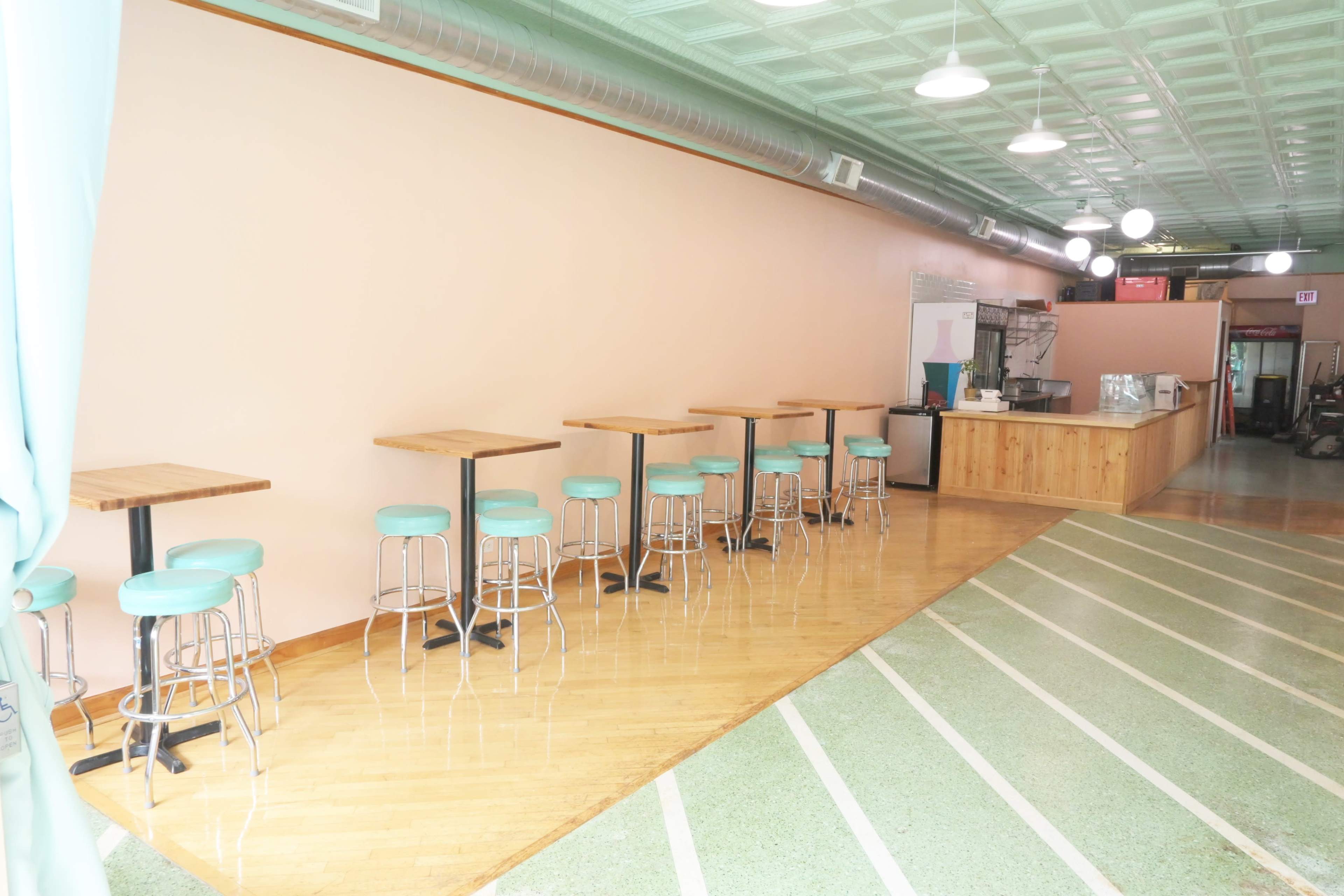 A cafe interior with a row of wooden tables and teal bar stools against a light-colored wall.