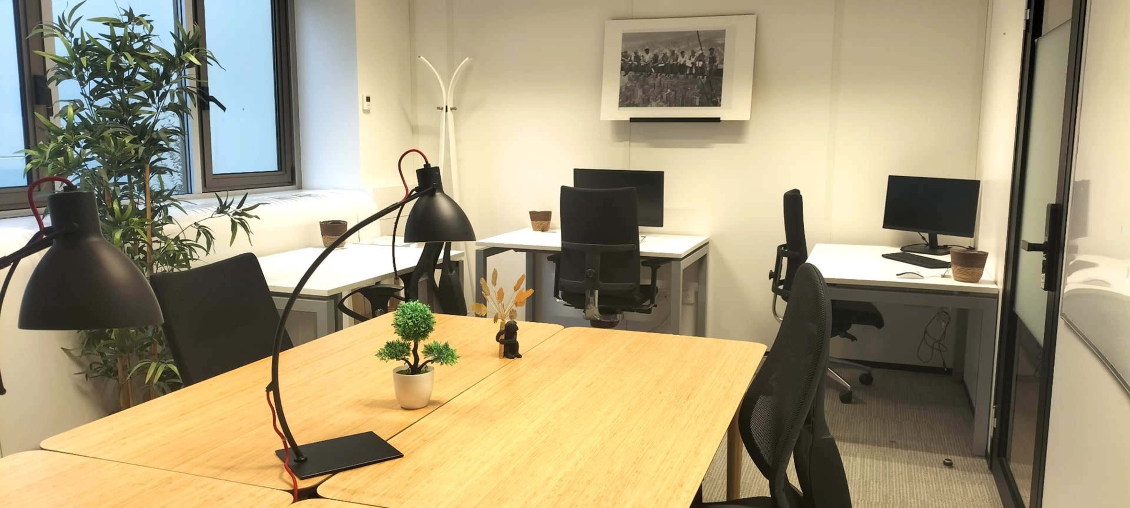 The image shows a modern office space with several desks, chairs, and computer monitors, illuminated by natural light from the windows.