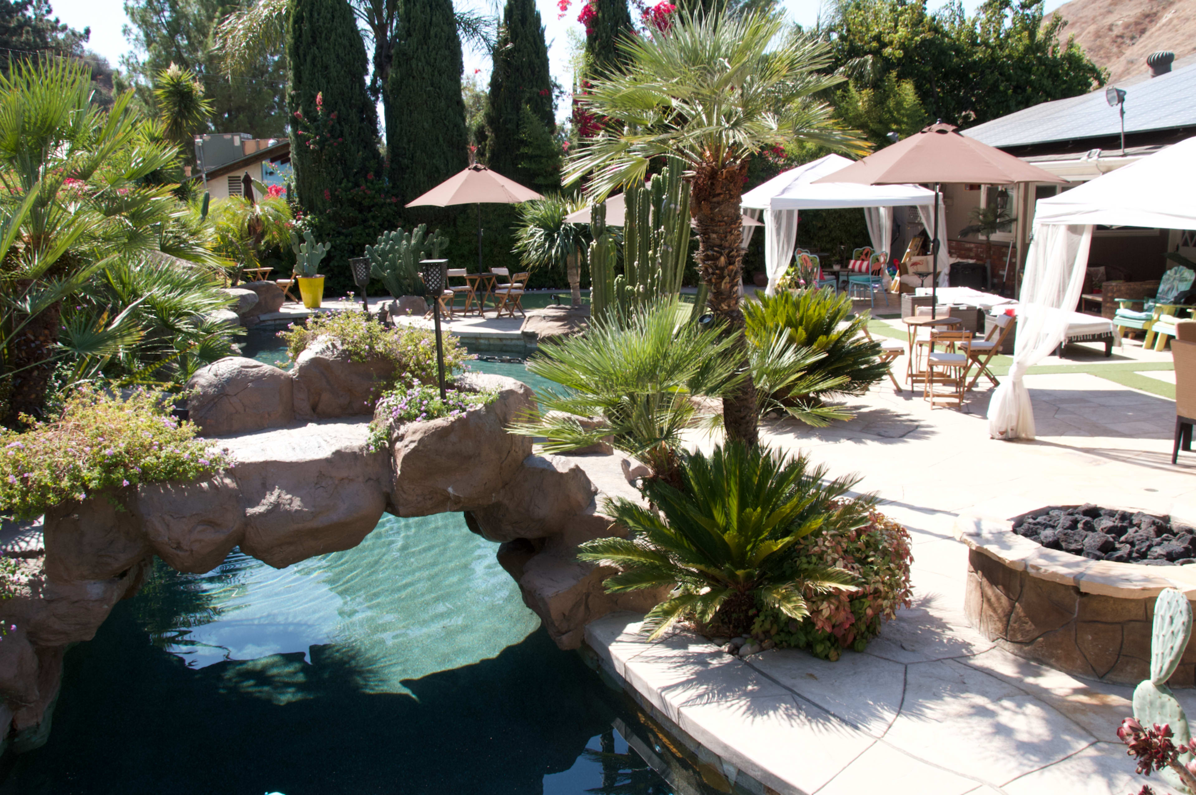 A landscaped backyard pool area with palm trees, a rock formation, and shaded seating under umbrellas.