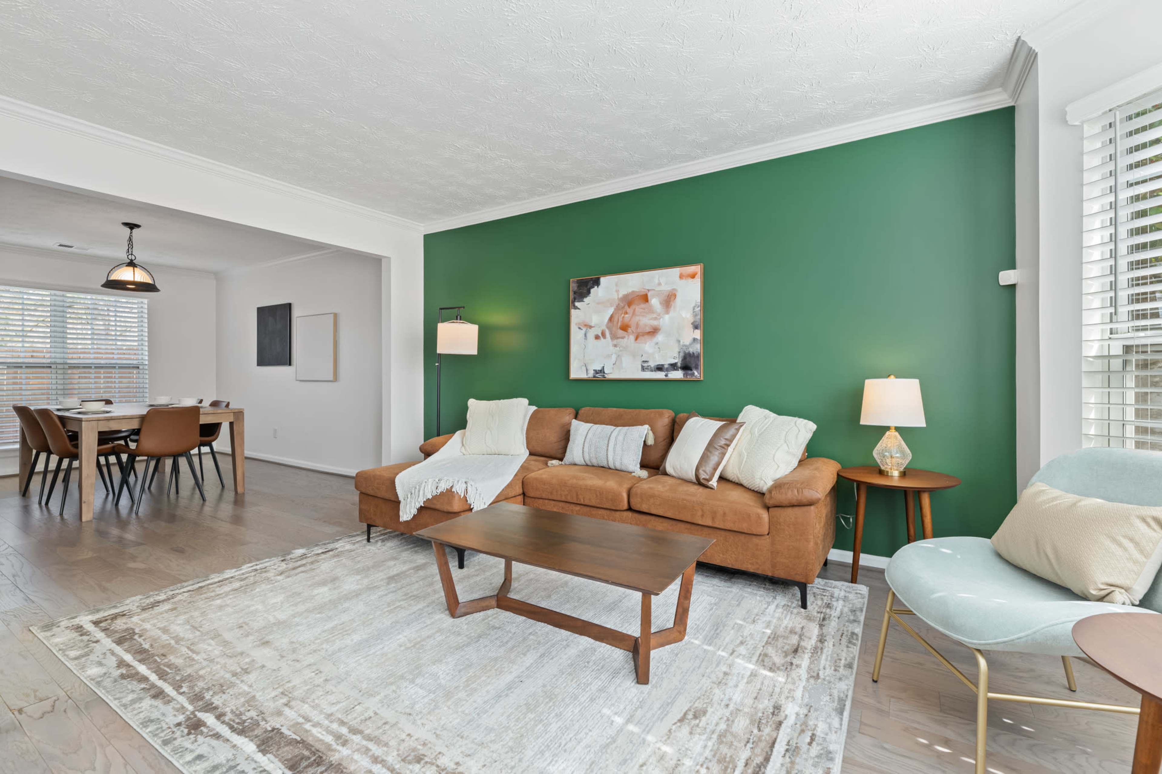 A living room with a brown sofa, a wooden coffee table, and a green accent wall, leading into a dining area with a table and chairs.