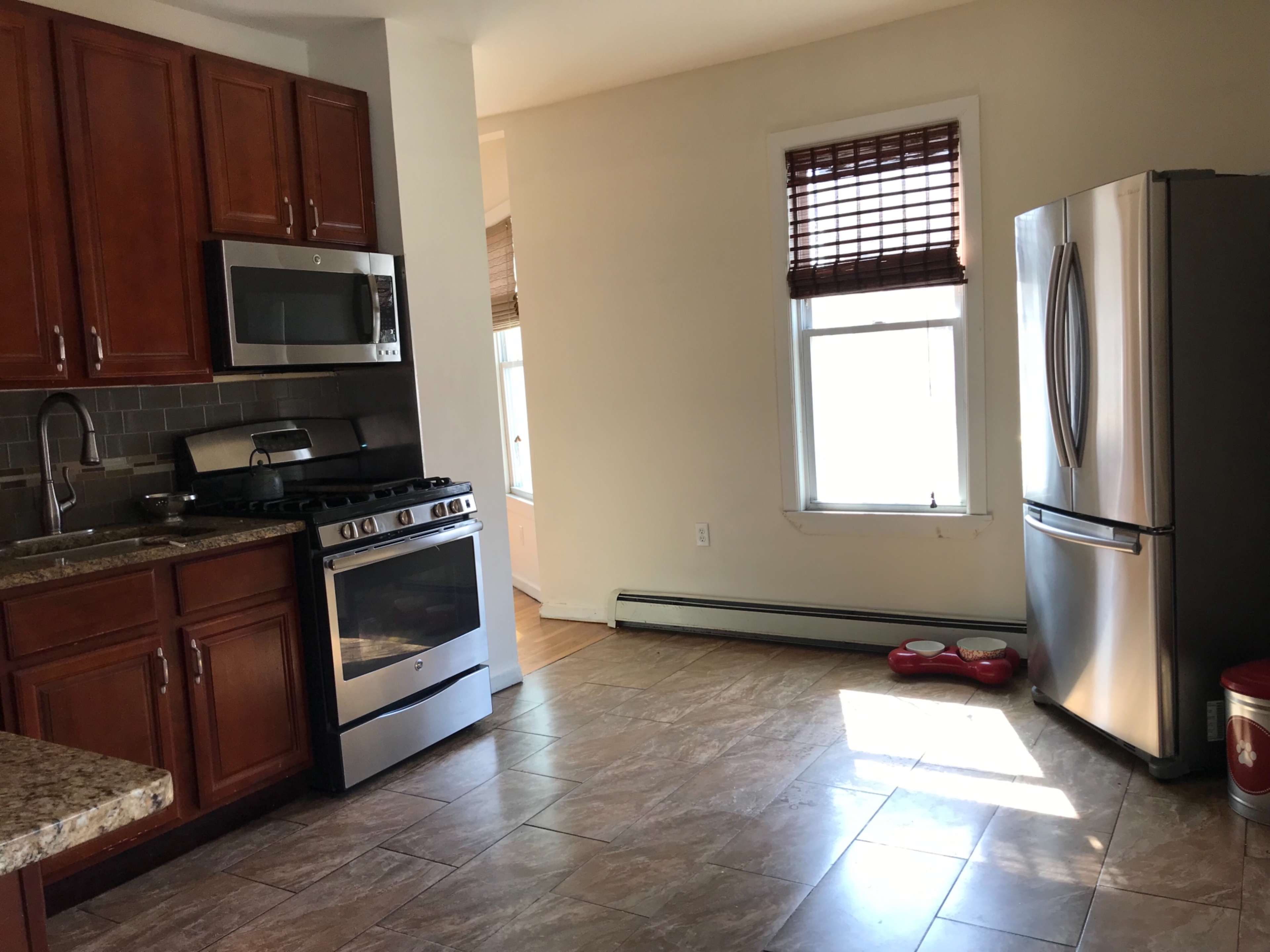 The image shows a kitchen area with wooden cabinets, a stainless steel refrigerator, and a gas stove, featuring tiled flooring and natural light coming through a window.