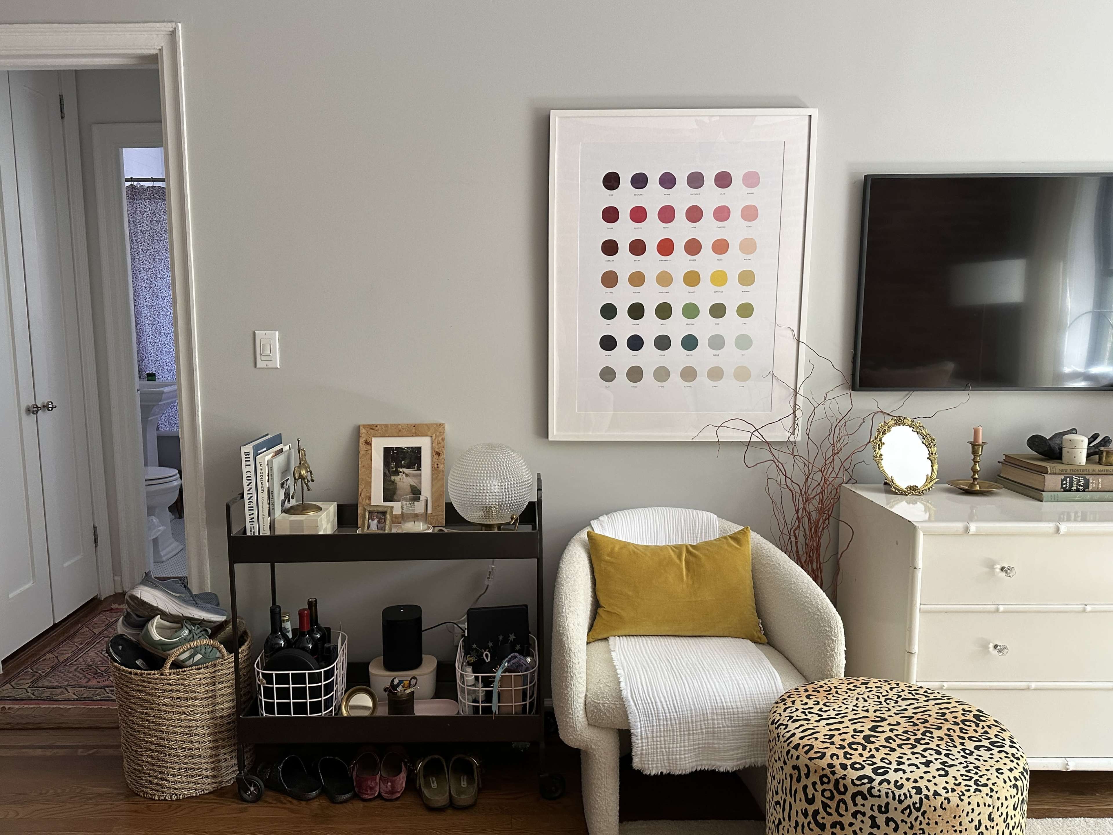 A living room with a decorative cart holding various items, a white chair, a leopard-print stool, a wall-mounted TV, and a colorful art print above a dresser.