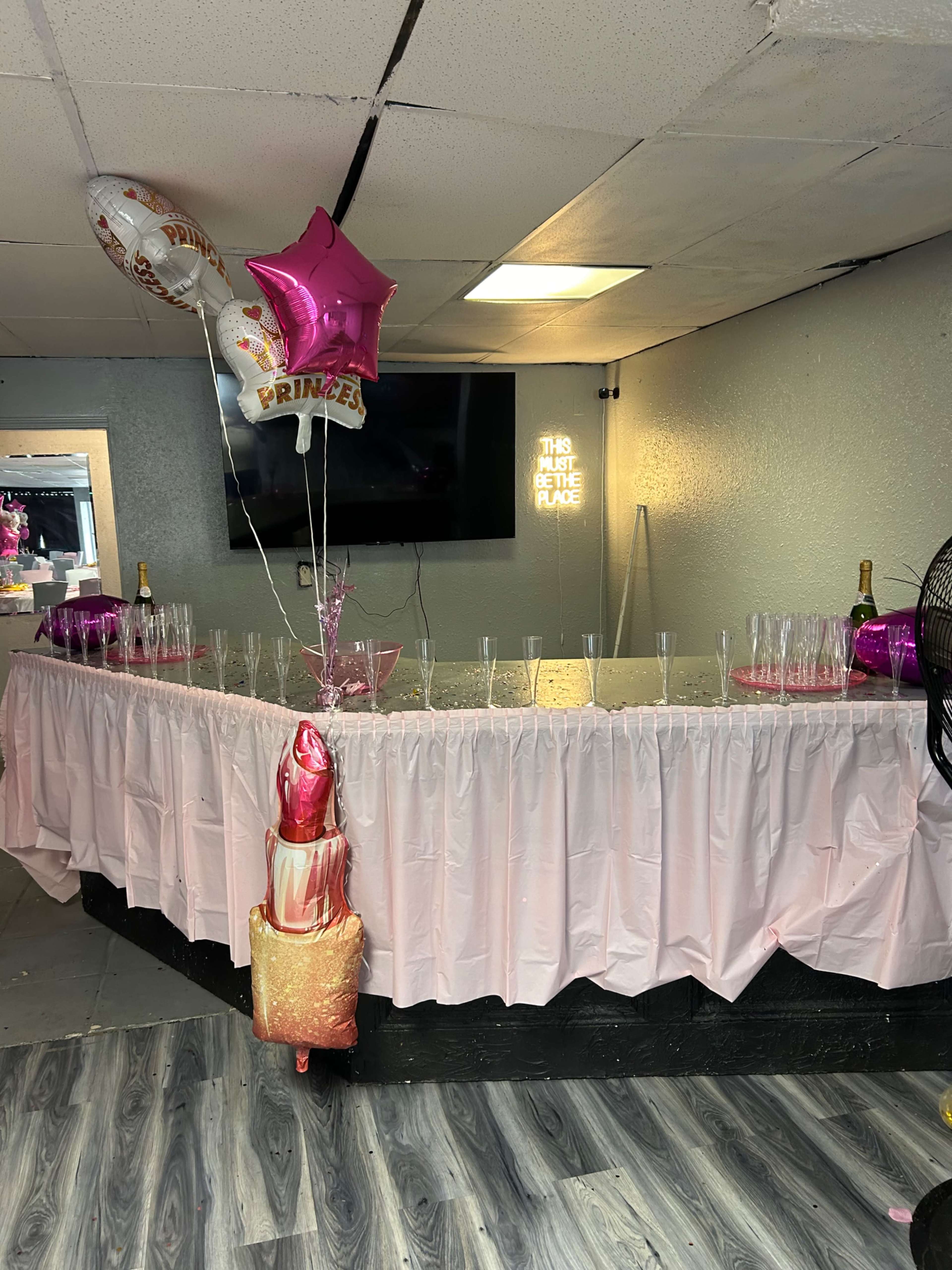 The image shows a party setup with a pink table skirt, multiple champagne glasses, balloons, and a decorative cake prop in a room with a television.