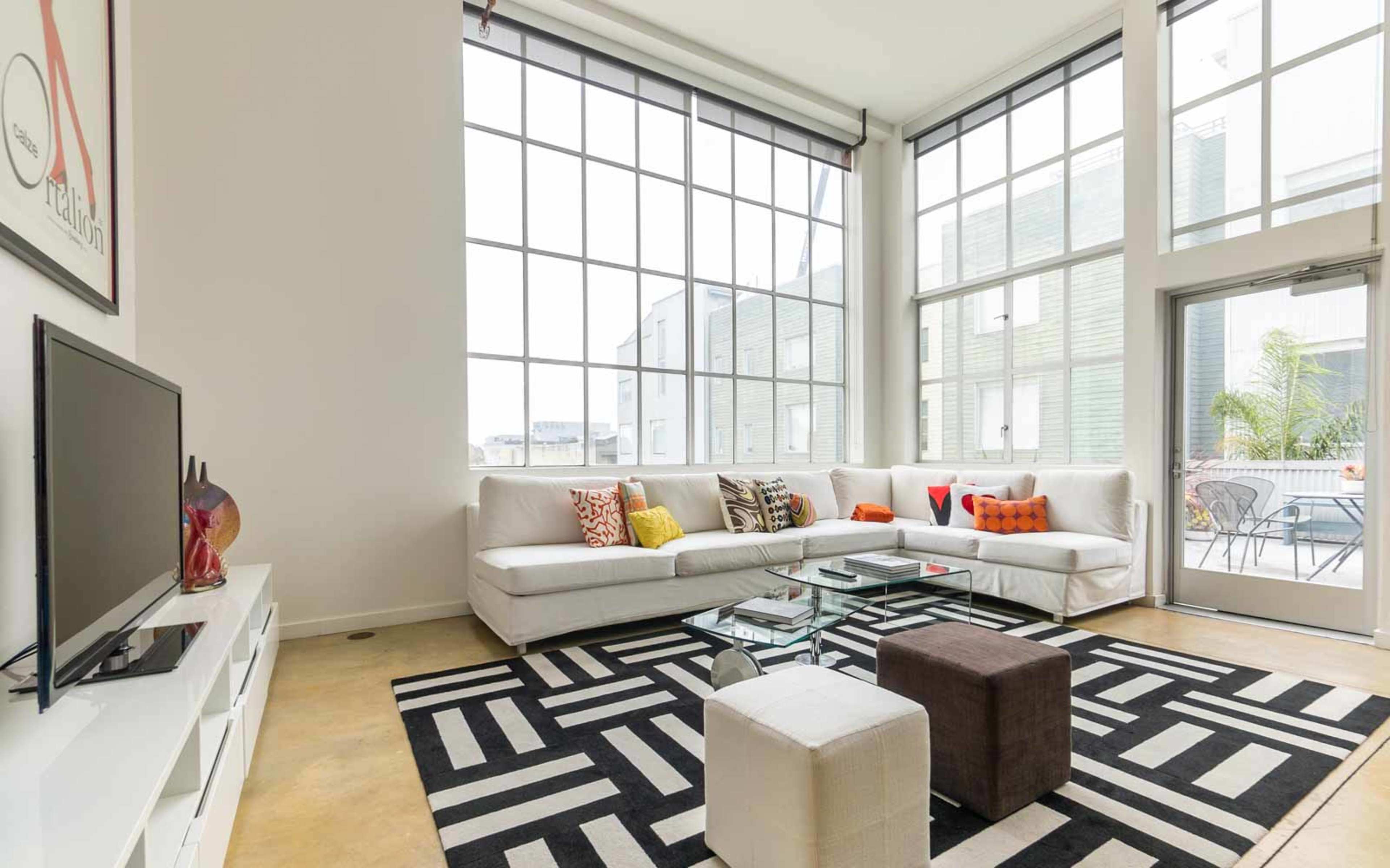 A modern living room features large windows, a sectional sofa with colorful pillows, a glass coffee table, and a geometric area rug.