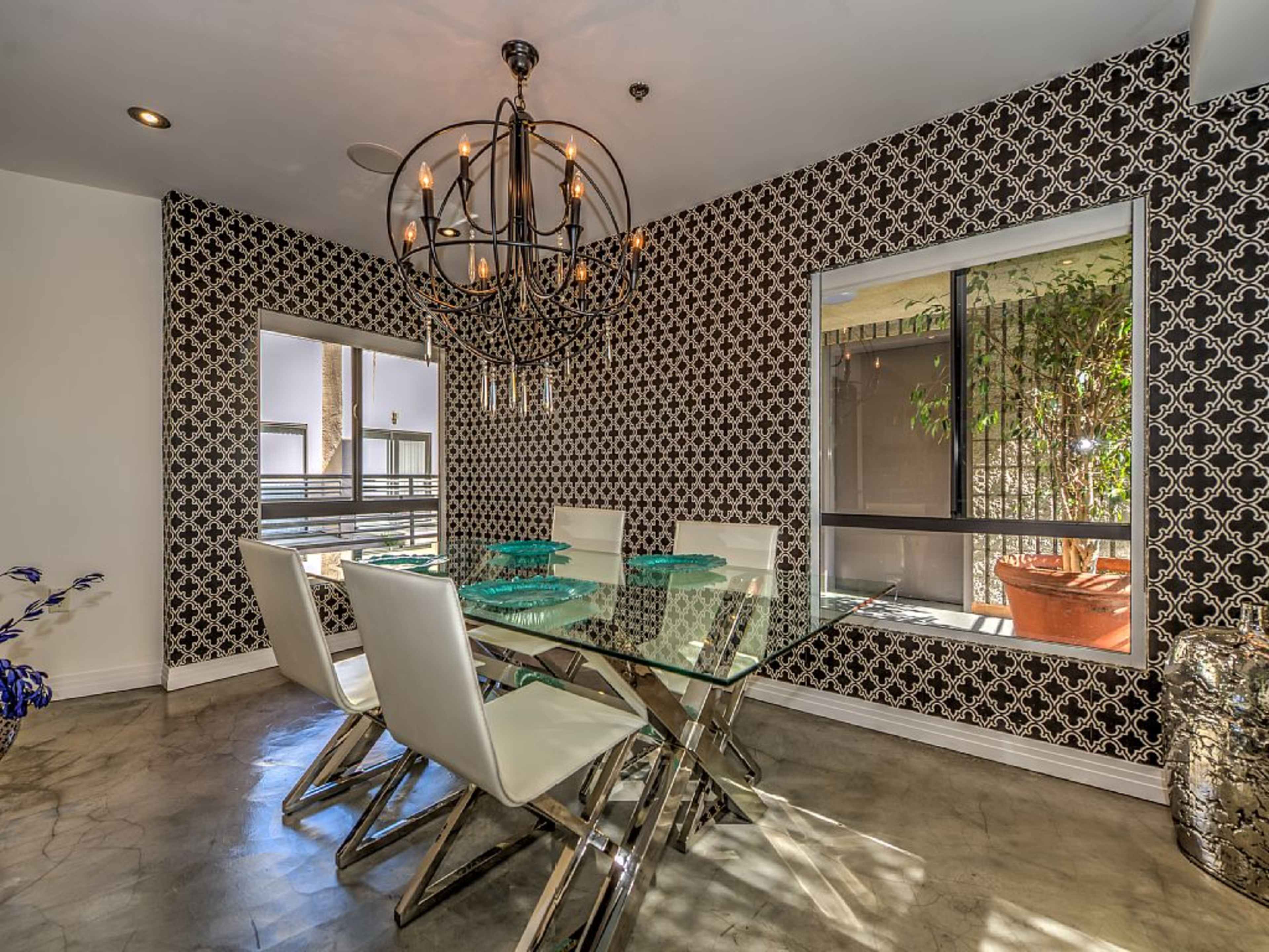 A dining area with a glass table surrounded by white chairs, set against a patterned black-and-white wall and featuring two windows with plants outside.