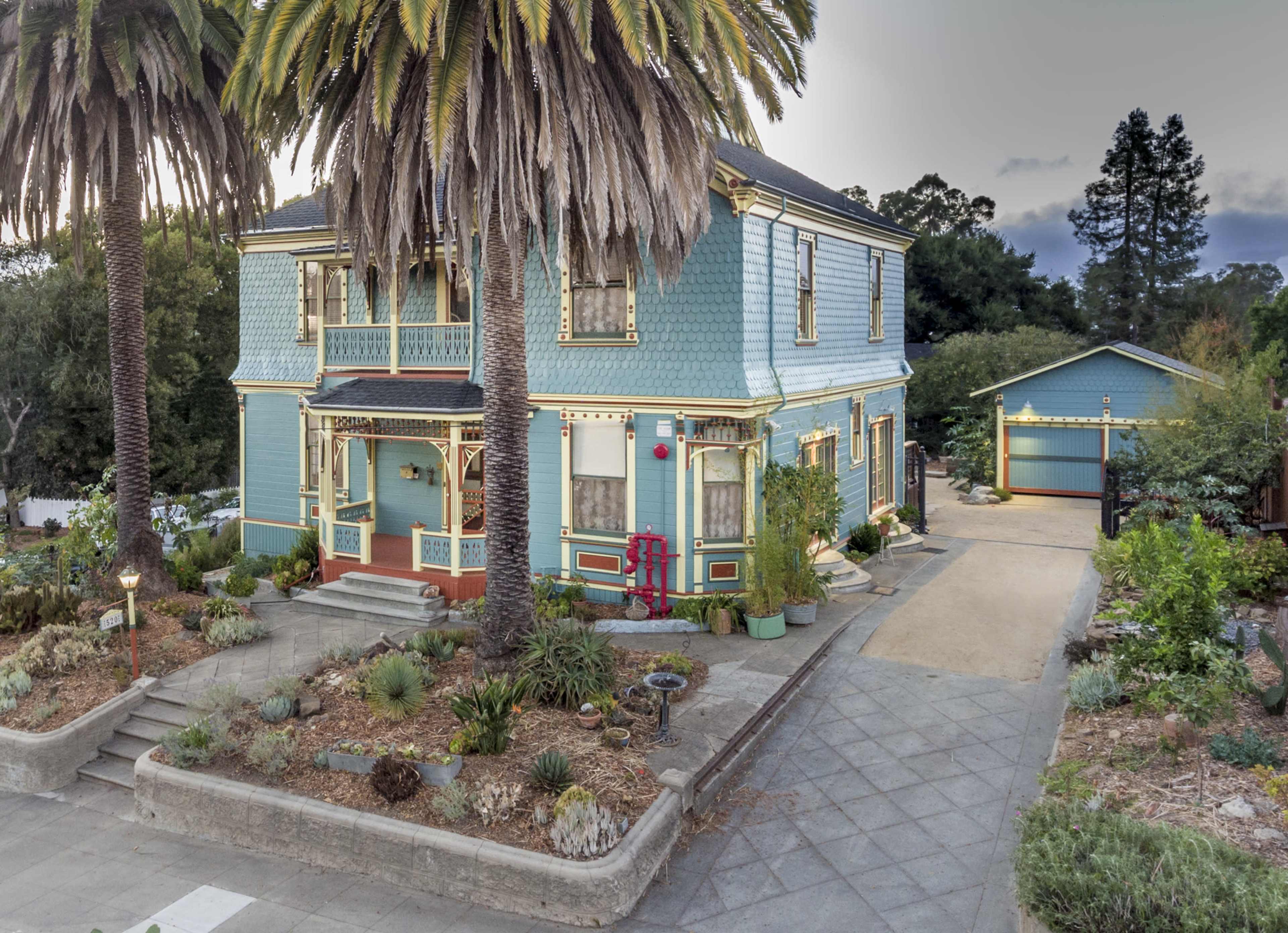 A two-story blue Victorian house with palm trees and a separate garage is surrounded by landscaped gardens.