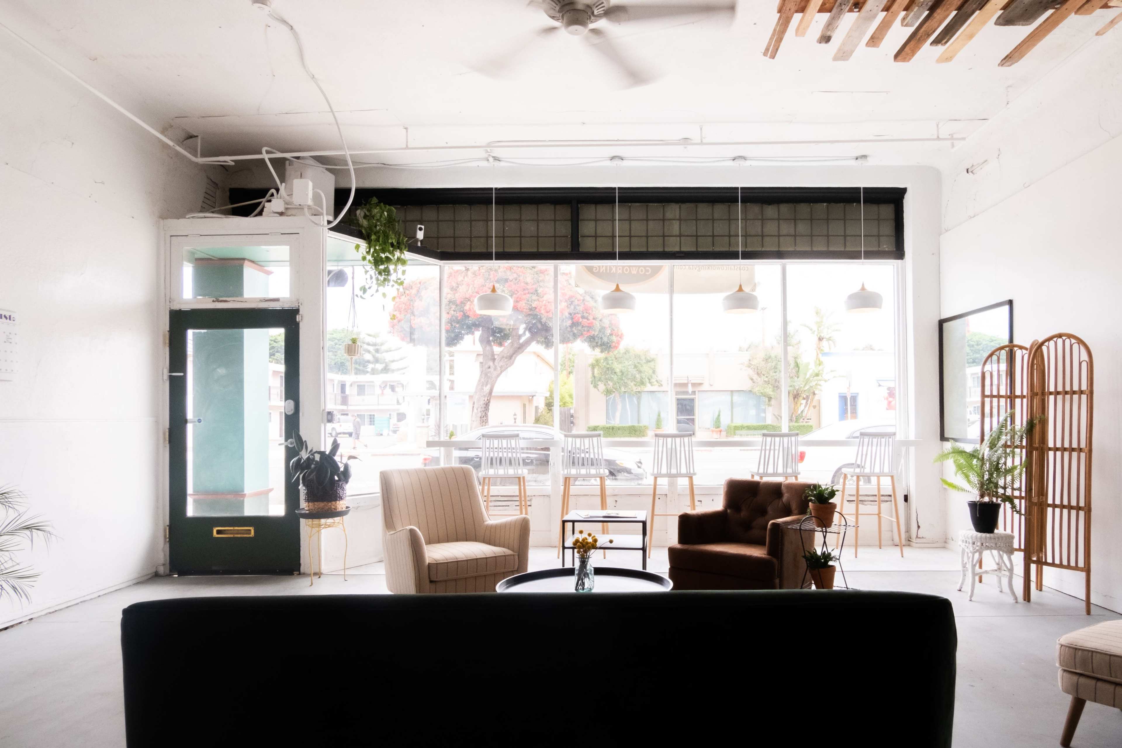 A cozy interior with a mix of seating options including armchairs and a sofa, alongside large windows offering a view of an outdoor area with tables and greenery.