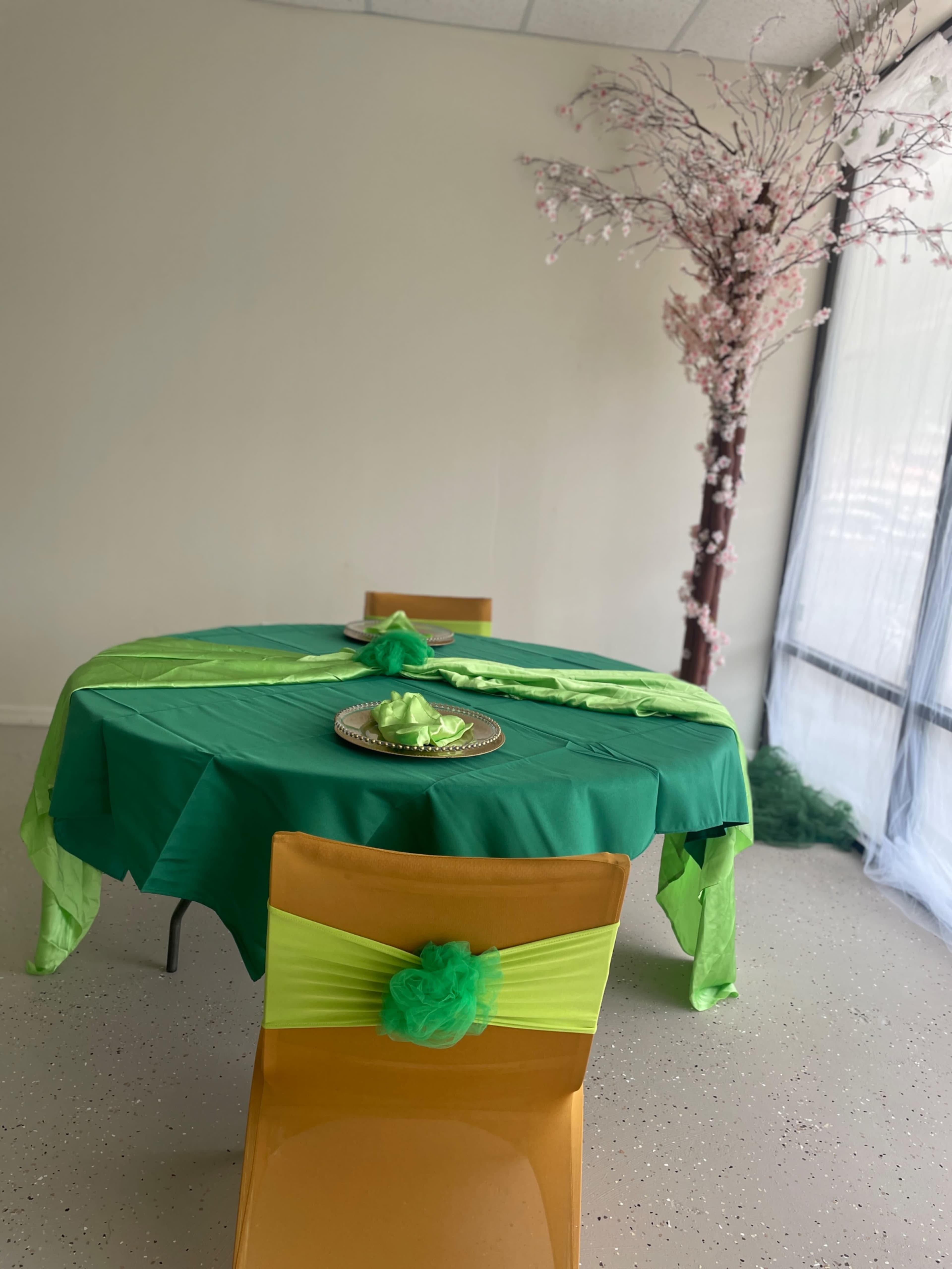 A round table covered with a green tablecloth is set with matching decorations, surrounded by chairs adorned with green sashes, next to a flowering tree in a bright interior space.